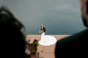 Bride and groom embrace on a wooden dock by the water, her white gown trailing elegantly, viewed from above.