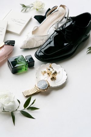 Elegant wedding flat lay with black patent shoes, white heels, gold watch, earrings, perfume bottles, and white roses on white background.