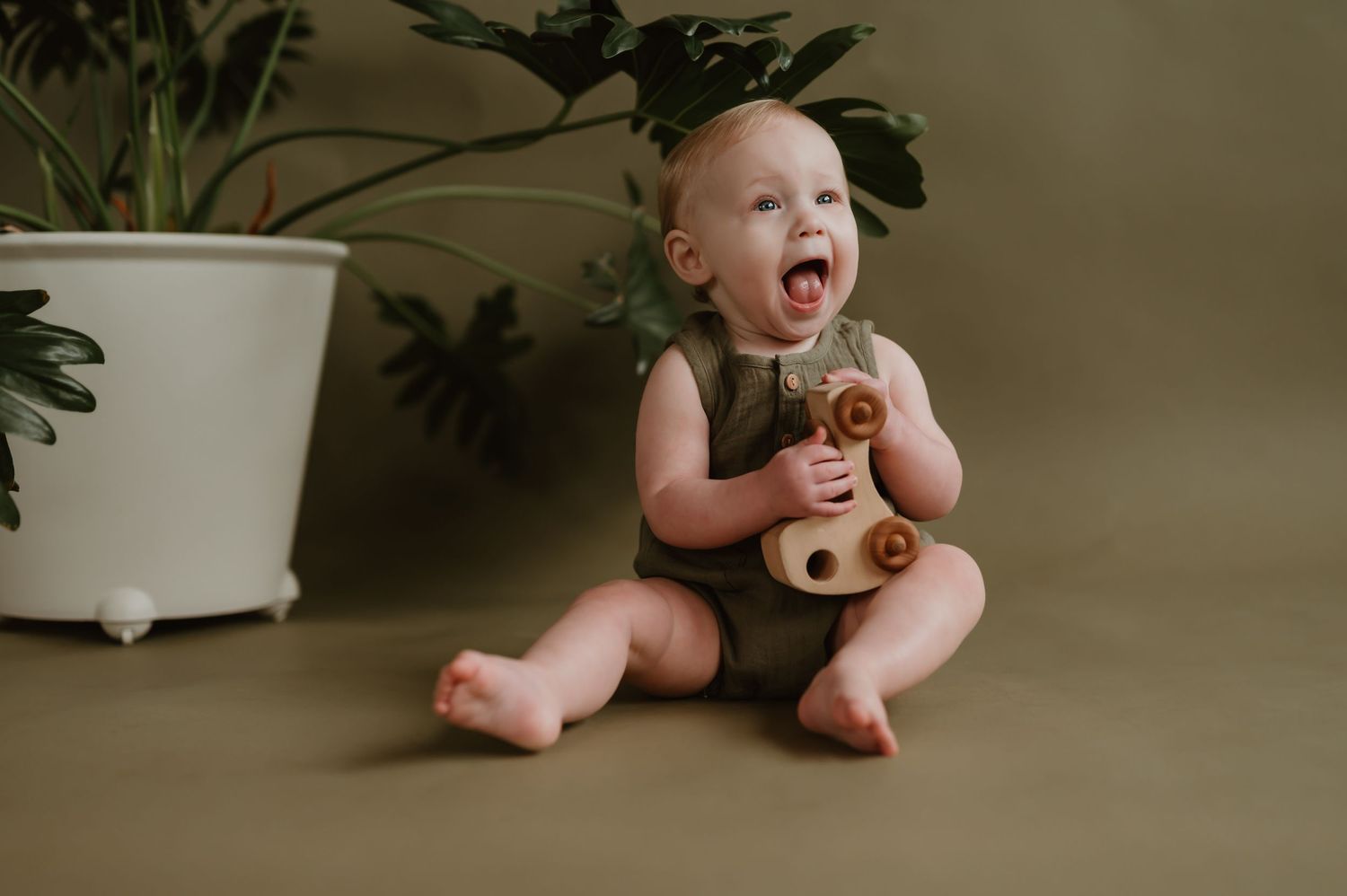 Happy baby in olive romper sits on tan floor, laughing and holding wooden toy beside a white potted plant.