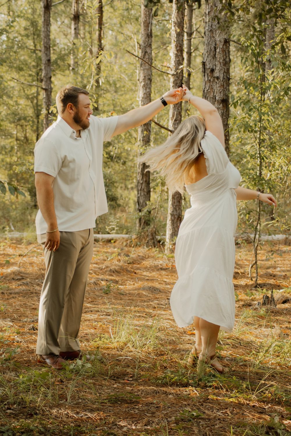 A couple dances together in a sunlit forest setting wearing white and earth-toned clothing.