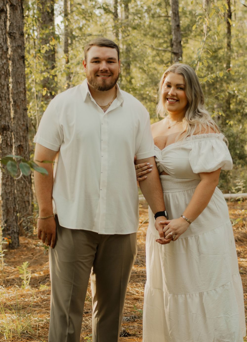 A couple in white and olive green outfits share intimate moments in a sunlit forest setting during a photo session.
