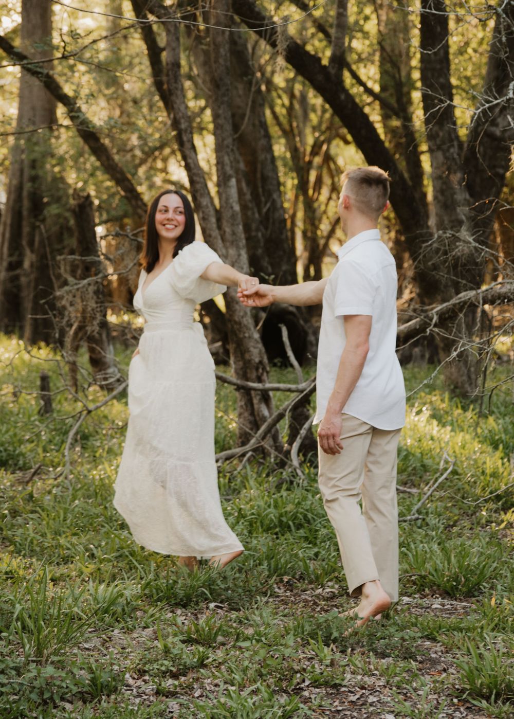 Happy couple dancing and holding hands in a sunlit forest clearing, woman wearing a flowing cream dress.