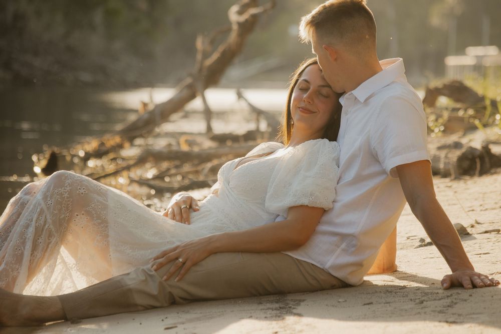Romantic couple resting together on a sandy beach at golden hour, woman in a white lace maternity gown.