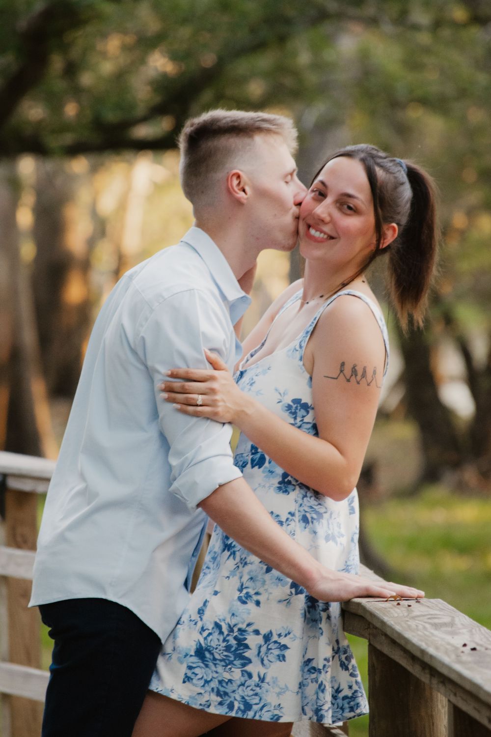 Man kissing smiling woman on the cheek as they embrace on a wooden bridge in an autumn park setting.