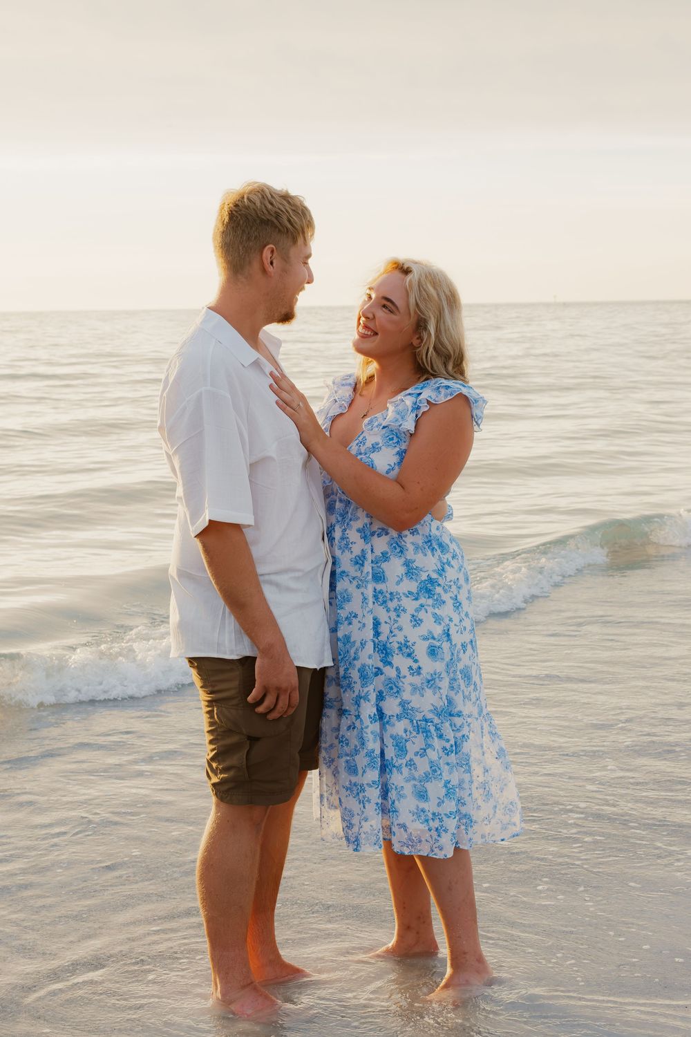 Couple standing barefoot in ocean waves at sunset, woman in blue floral dress smiling at man in white shirt.