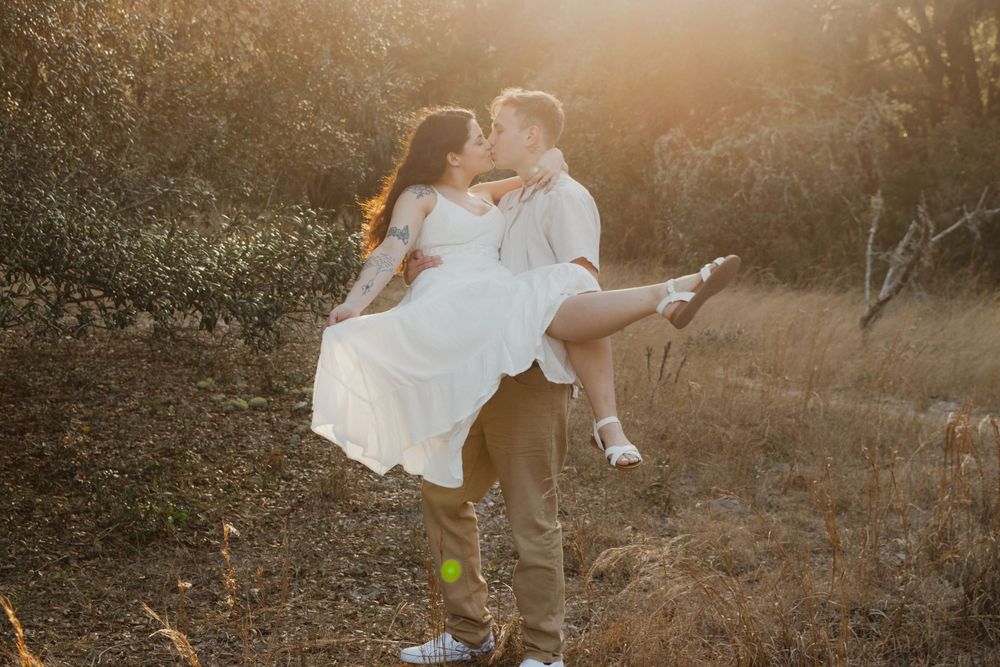 A couple in white and brown outfits embraces during a romantic sunset photoshoot in a golden field.