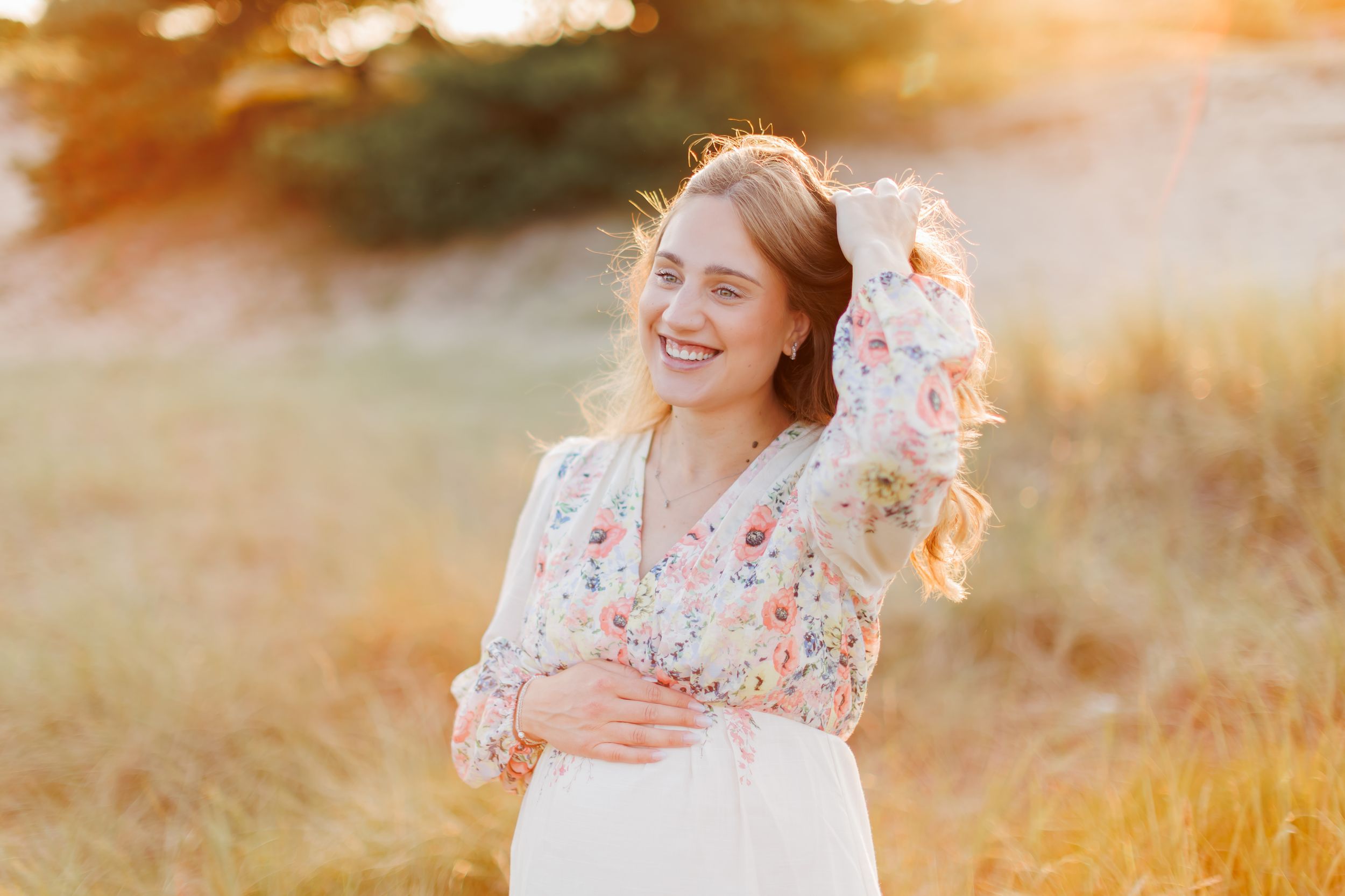 Young woman in floral blouse smiling outdoors at golden hour sunset in a field.
