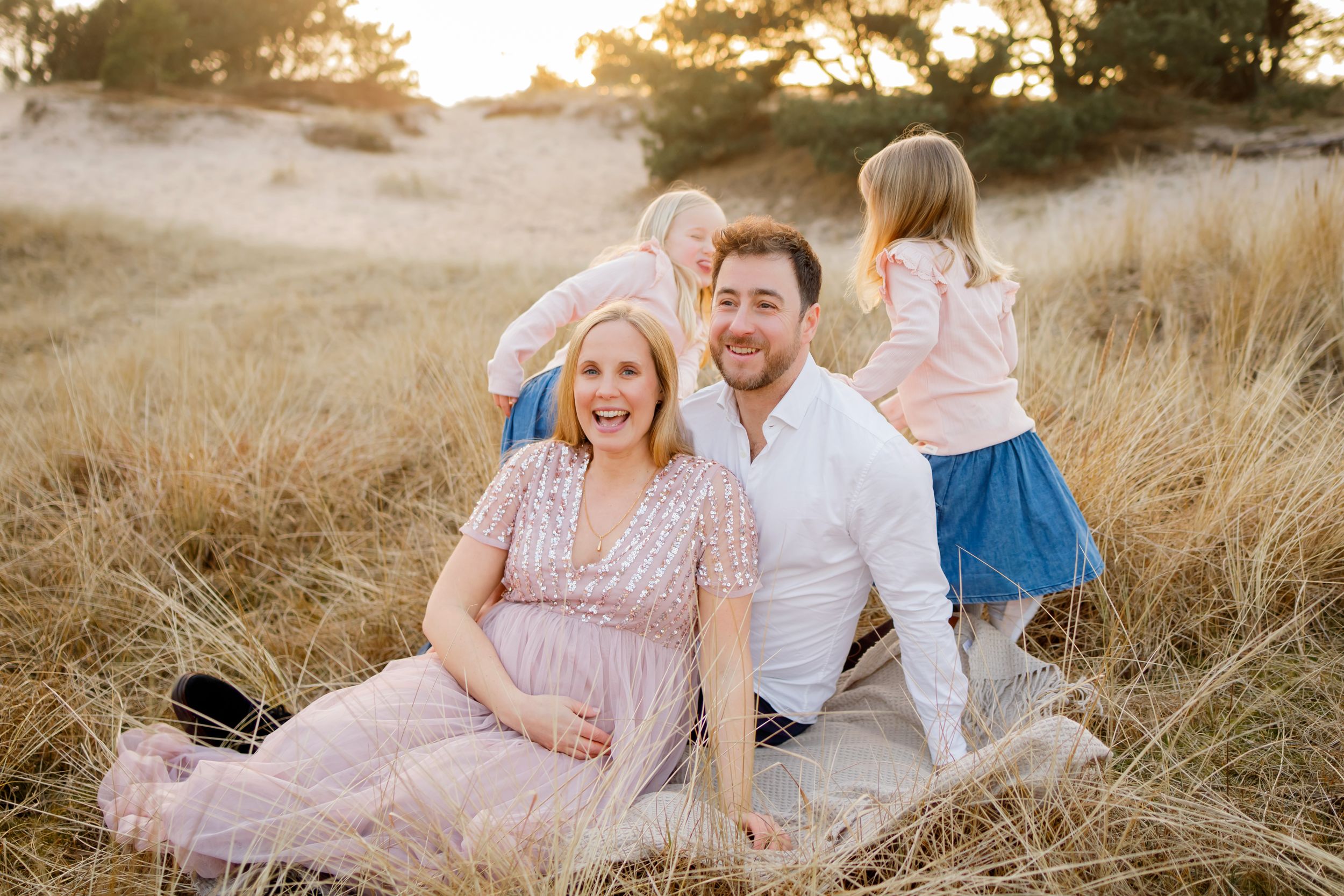 Pregnant woman and husband sit smiling in golden field with two young children climbing on their backs.