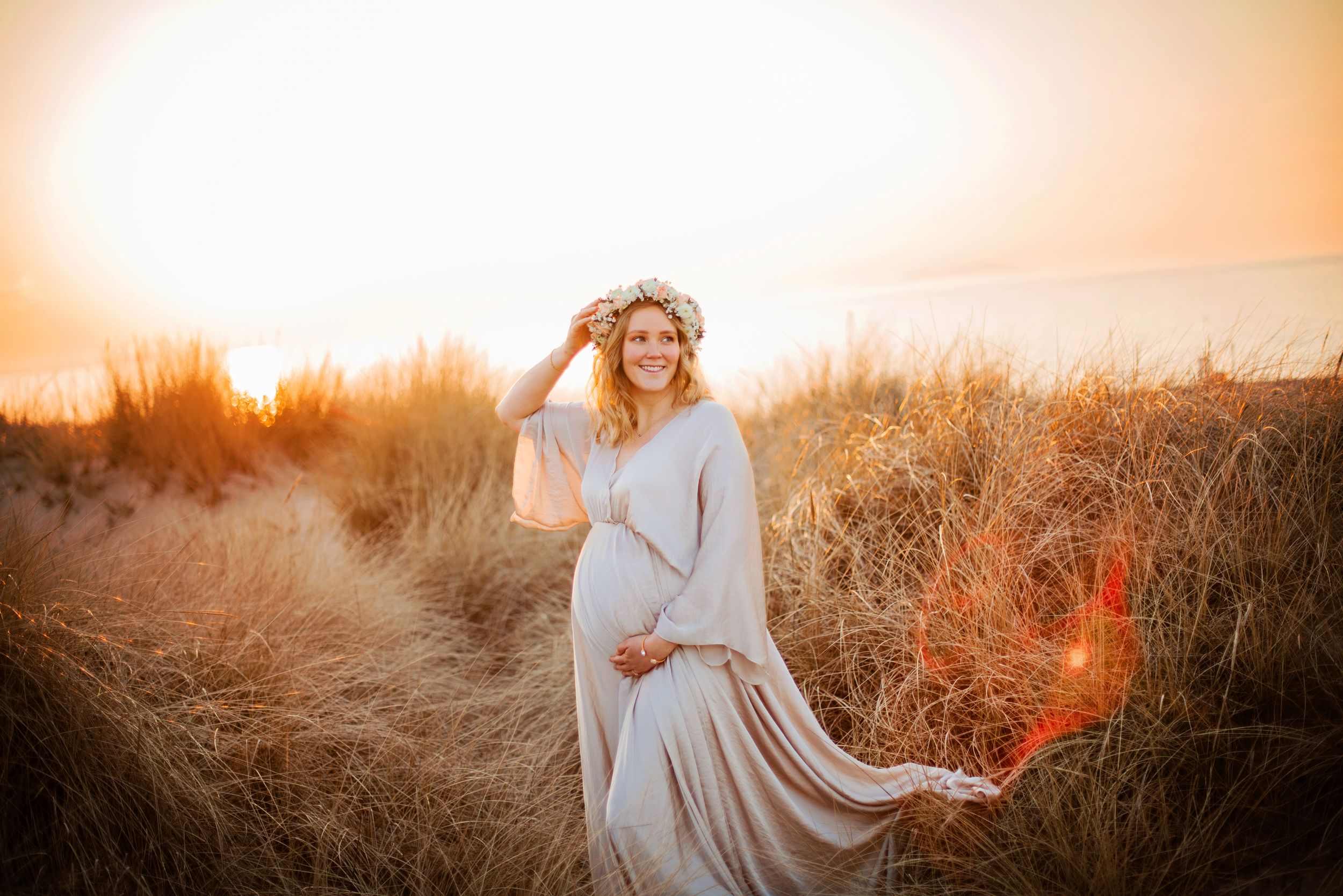 Pregnant woman in flowing beige gown and floral crown standing in golden grass field at sunset.