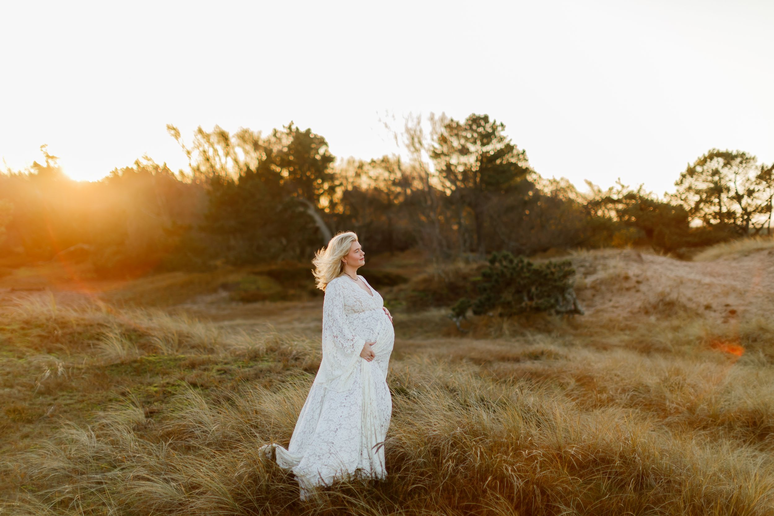 A person in a flowing white dress stands in a golden field at sunset with warm sunlight streaming through the trees.