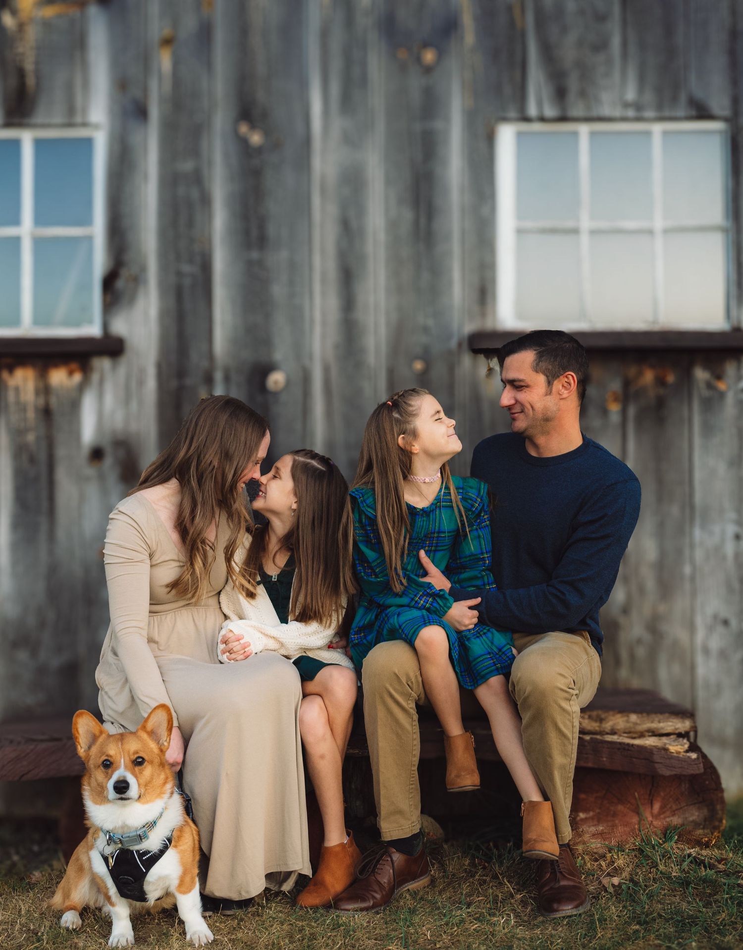Family sitting on rustic porch with corgi dog in front of weathered barn.