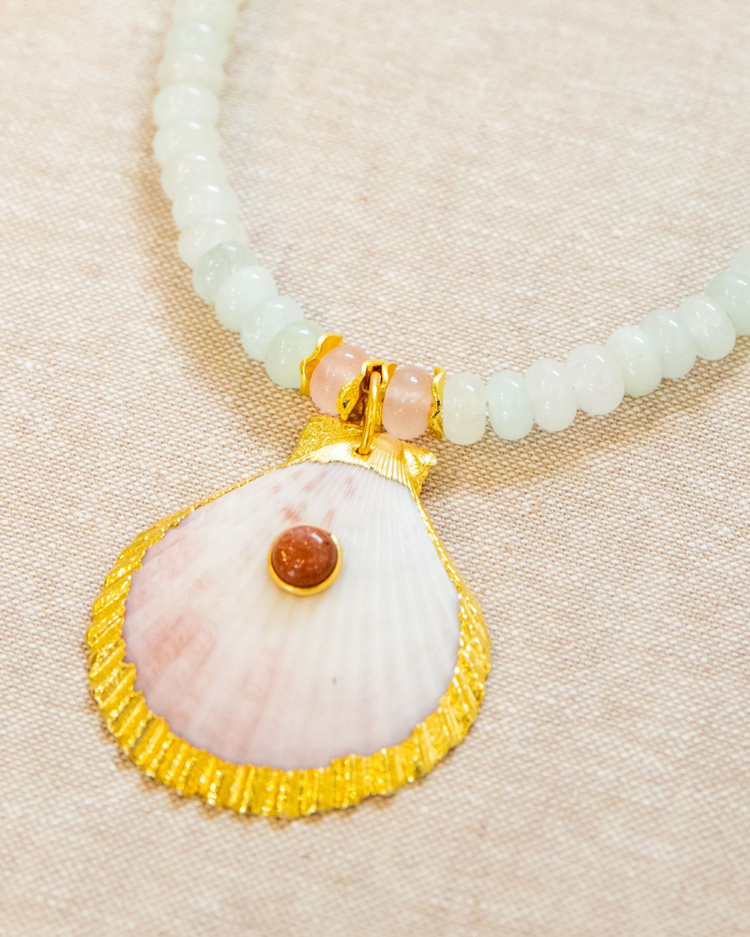 Elegant white beaded necklace with pink seashell pendant trimmed in gold displayed on neutral fabric.