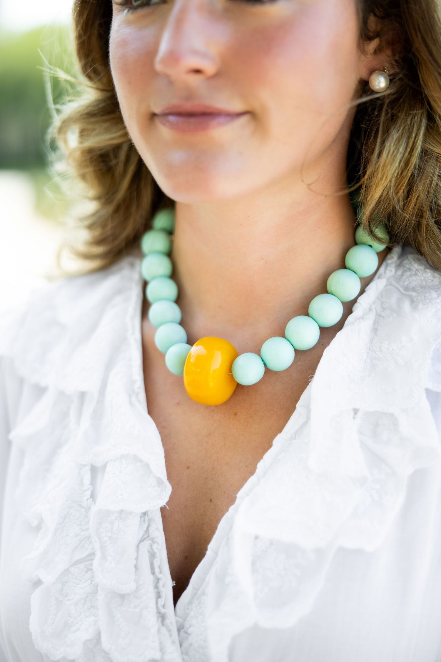 A statement necklace with mint blue beads and a bold yellow accent bead is shown against a white ruffled blouse.