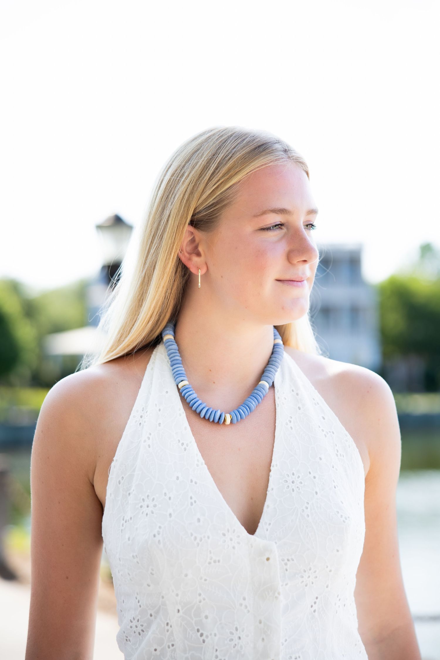 Woman in white eyelet dress wearing a blue beaded statement necklace against a waterfront backdrop.