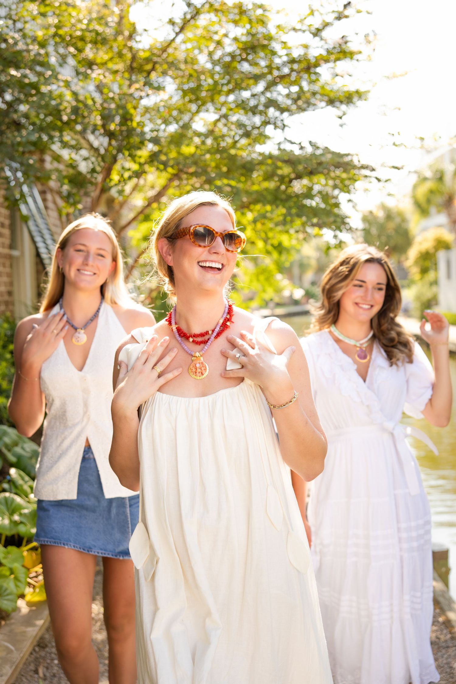 Three friends in white summer dresses laughing and enjoying a sunny day outdoors.