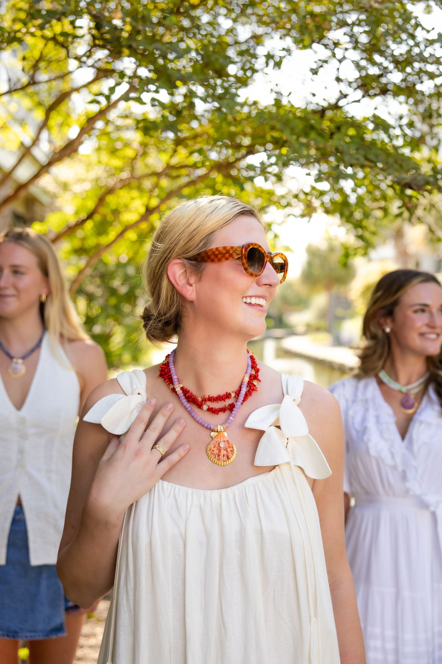 Group wearing white summer outfits and colorful accessories gathered outdoors under sunlit trees.