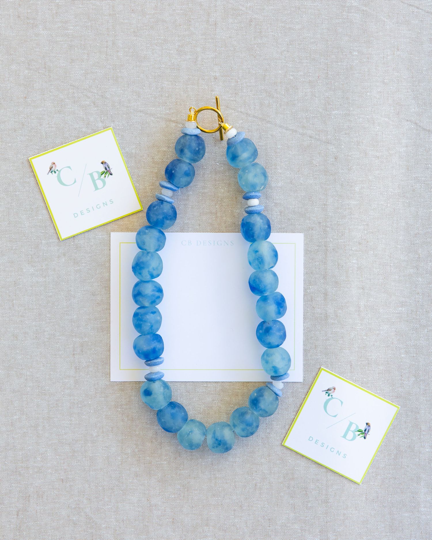 Four identical handmade blue glass bead bracelets displayed on a light gray fabric background with small white tags.