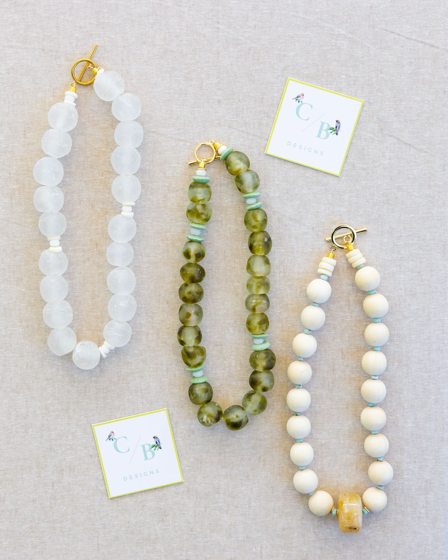 Collection of beaded necklaces featuring white, green and gold beads laid out on gray linen fabric.