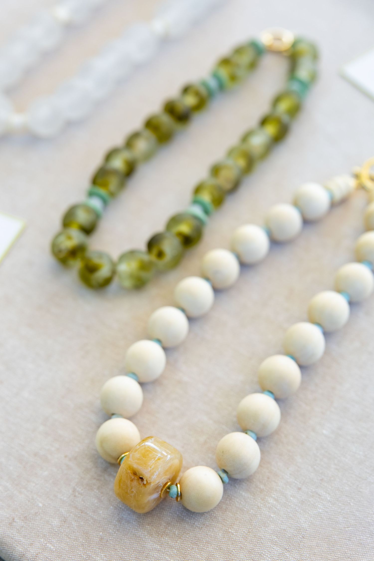 Two beaded necklaces displayed on light fabric, one with green glass beads and one with white spherical beads.