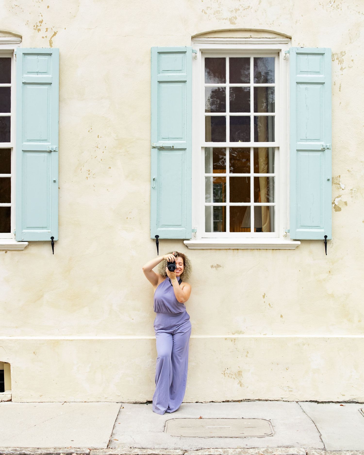 Photographer in purple jumpsuit taking photos against cream wall with blue shutters.
