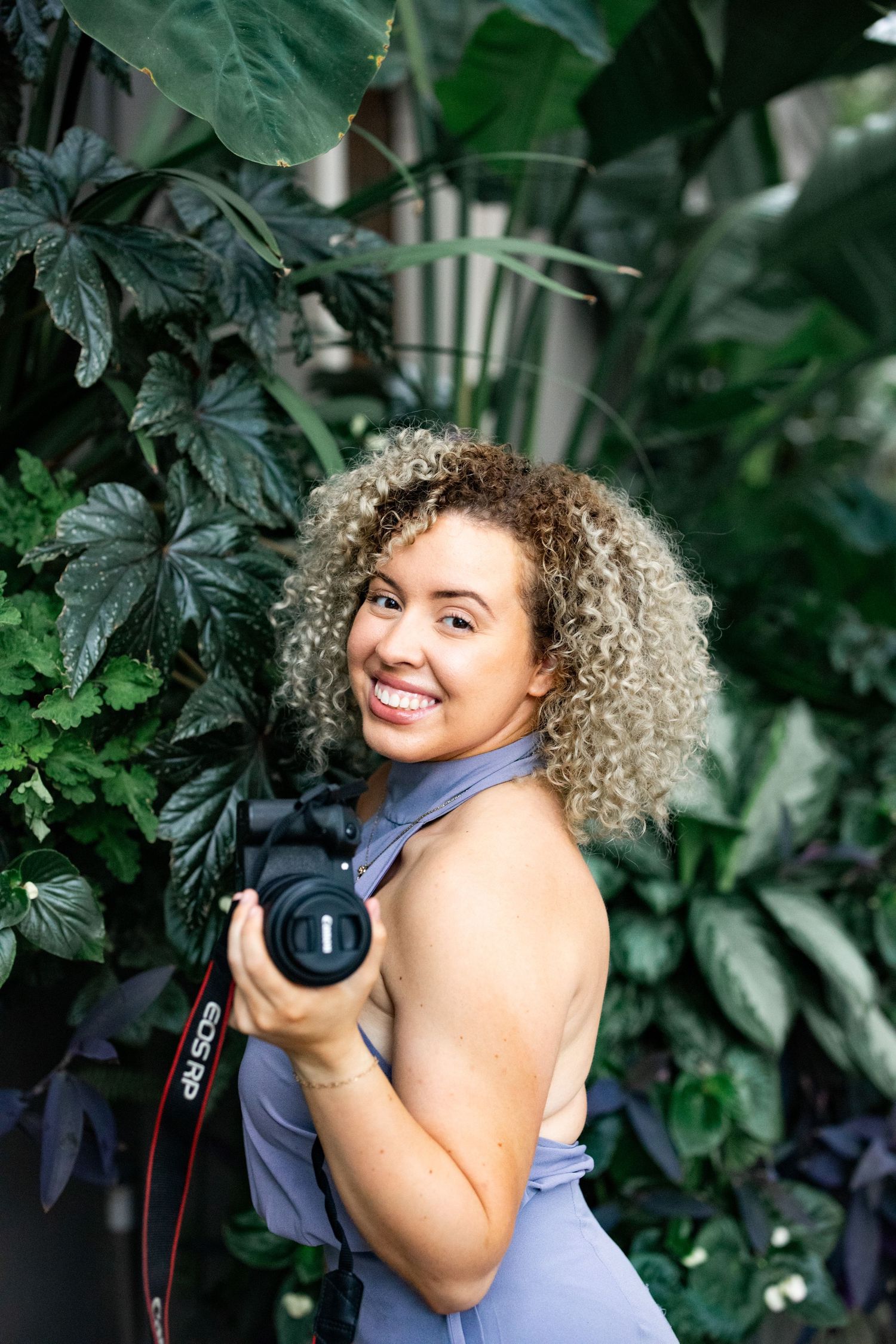 Photographer in purple outfit smiling with camera among tropical green foliage.