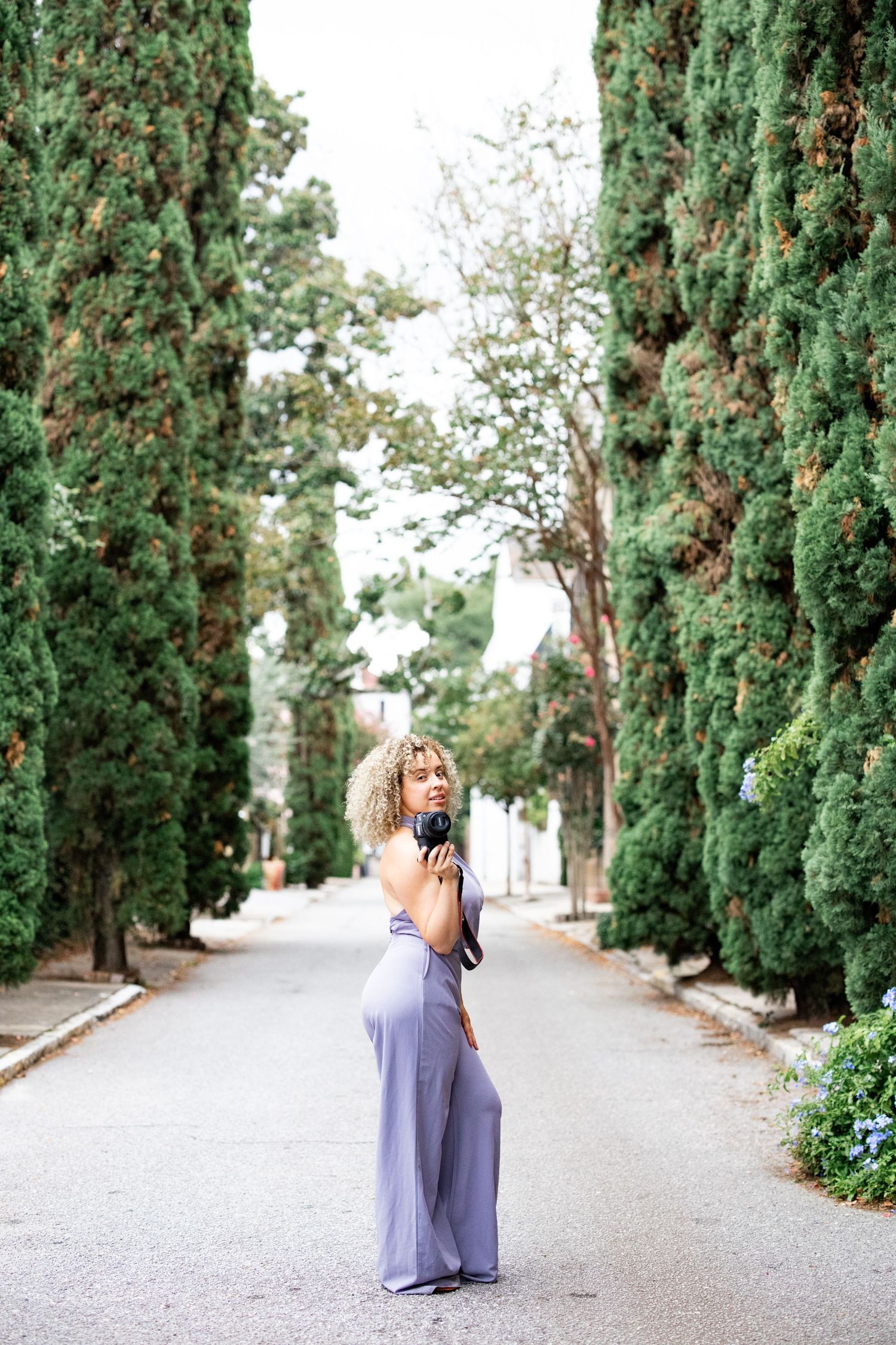 Photographer in lavender jumpsuit poses with camera on tree-lined path.