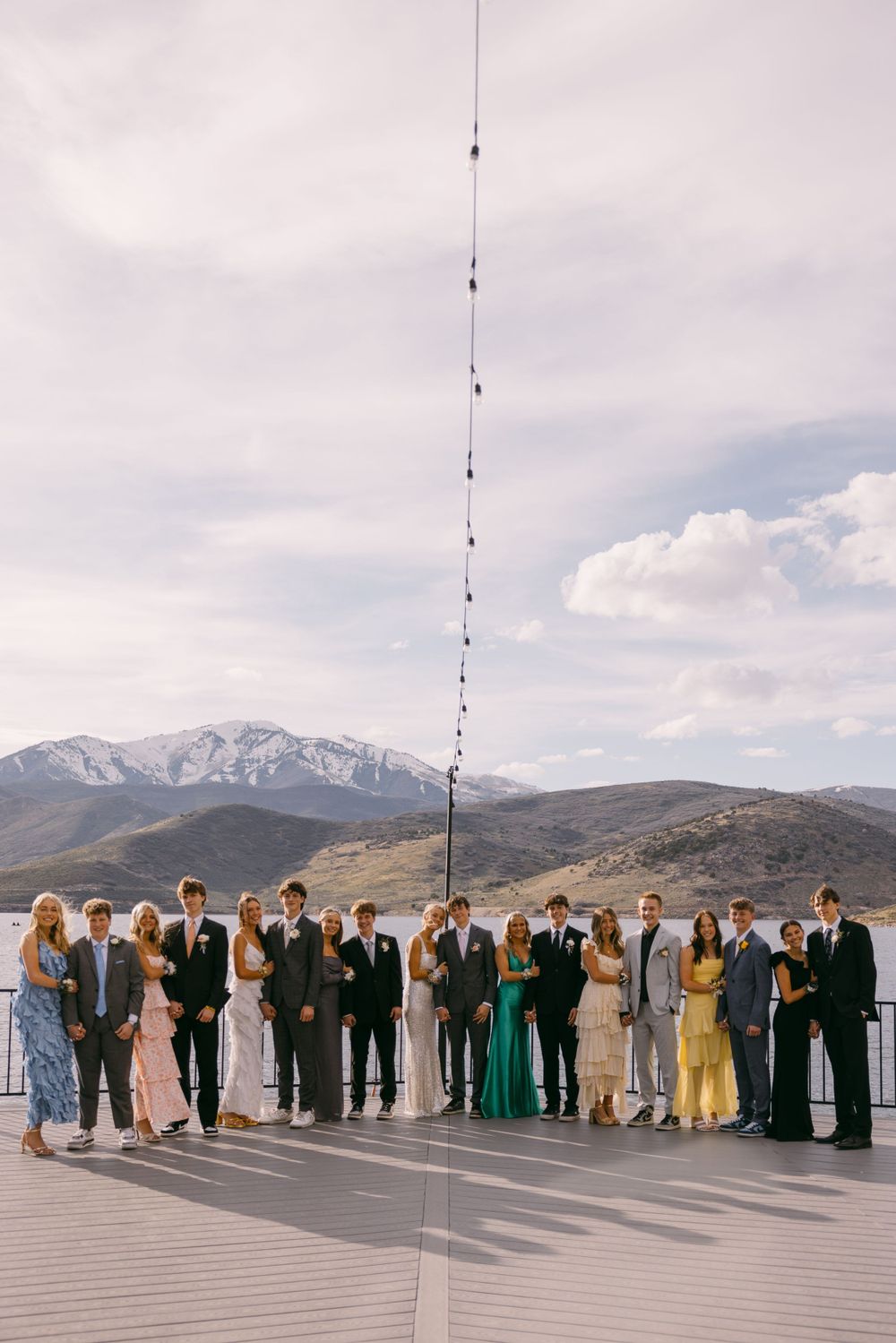 Wedding party in formal attire poses under string lights on a deck with mountain and lake views.
