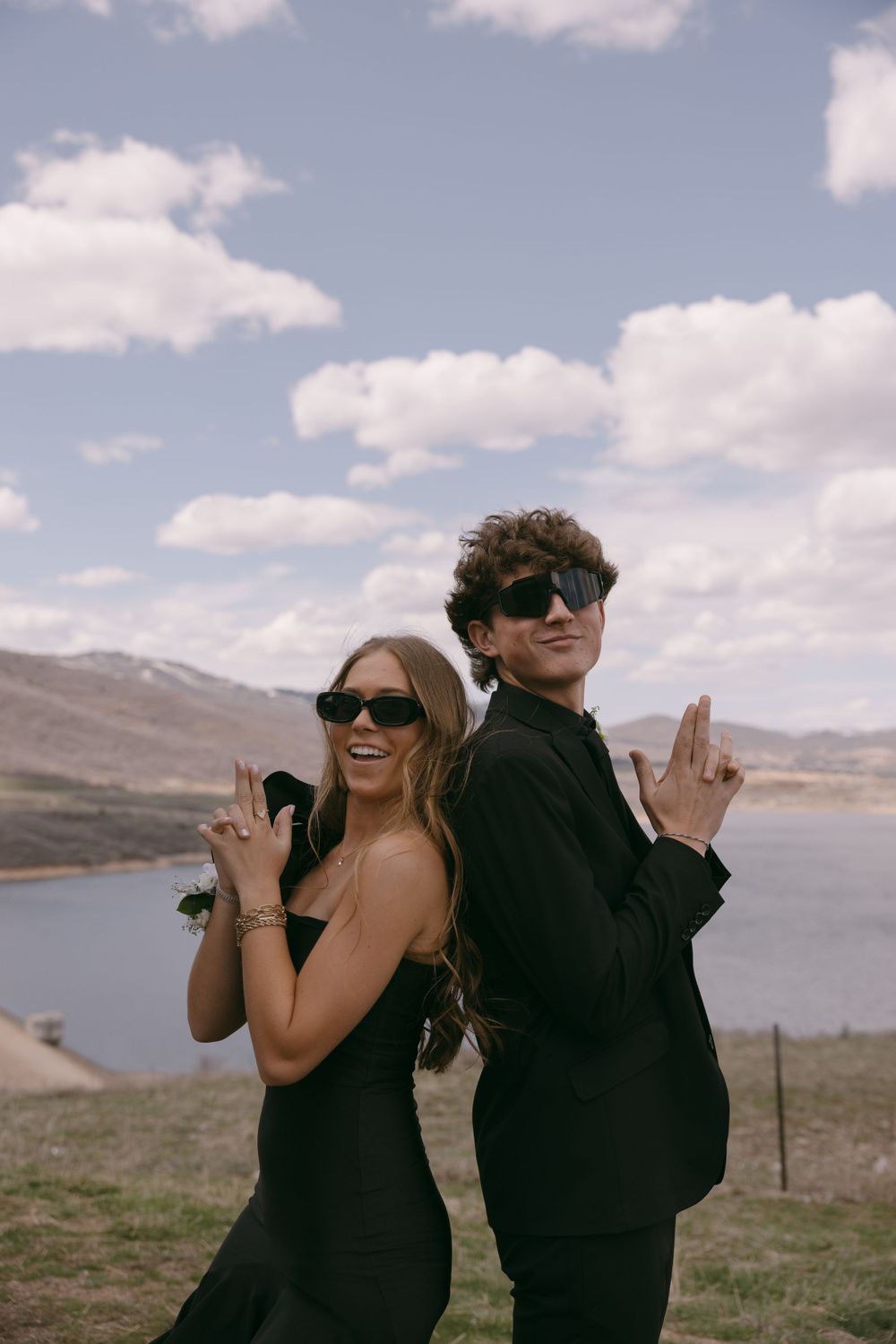 Two people in black outfits strike playful poses against a mountain landscape.