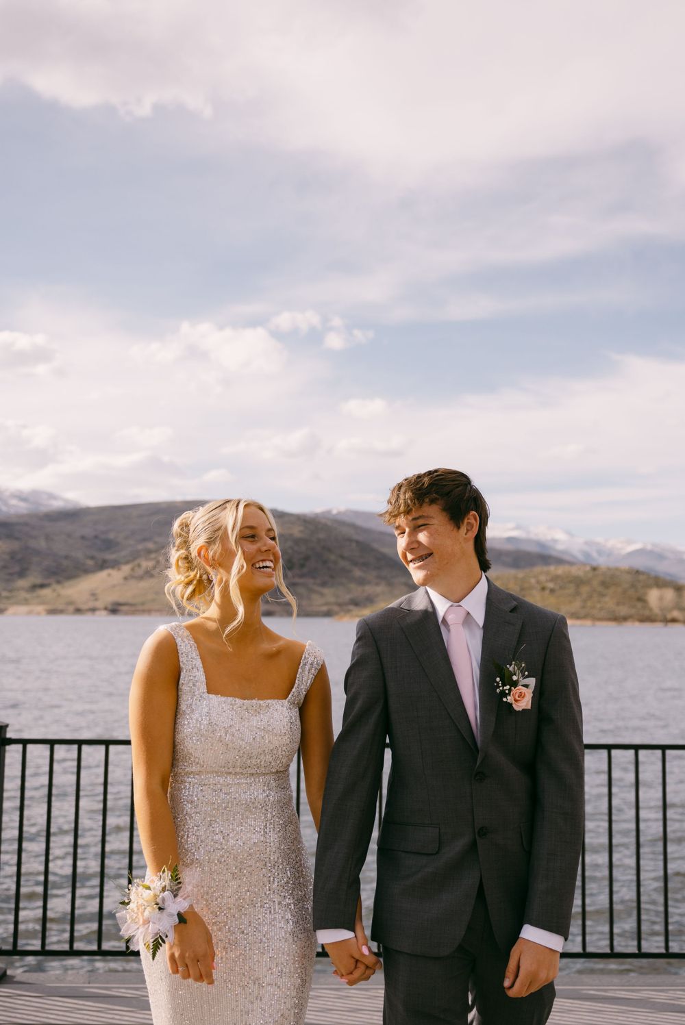 A couple in formal attire shares a happy moment on a deck overlooking a mountain lake.