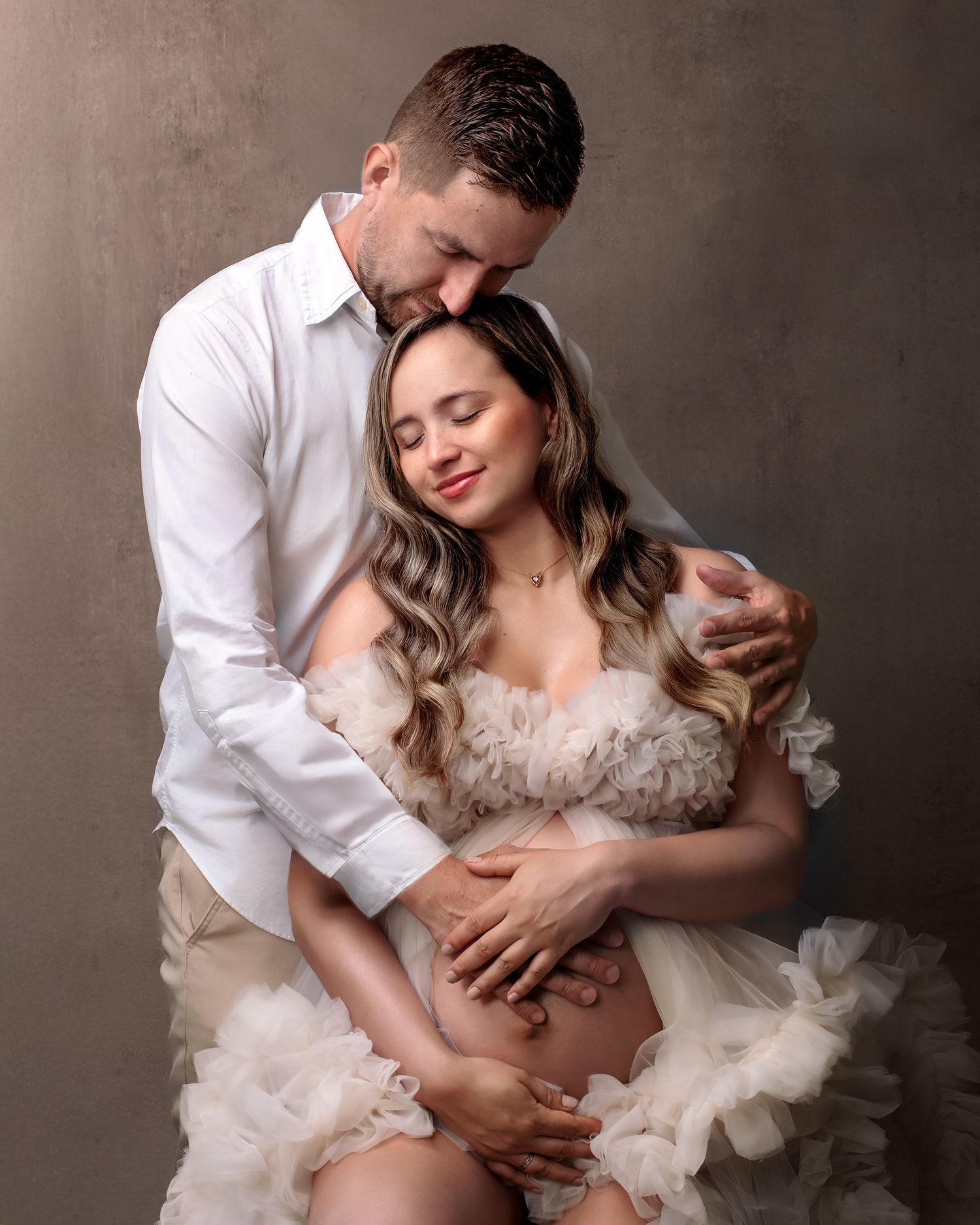 Fine art pregnancy photo: mum in a couture sits, with partner's hand on her belly and shoulder, elegant and supportive pose.