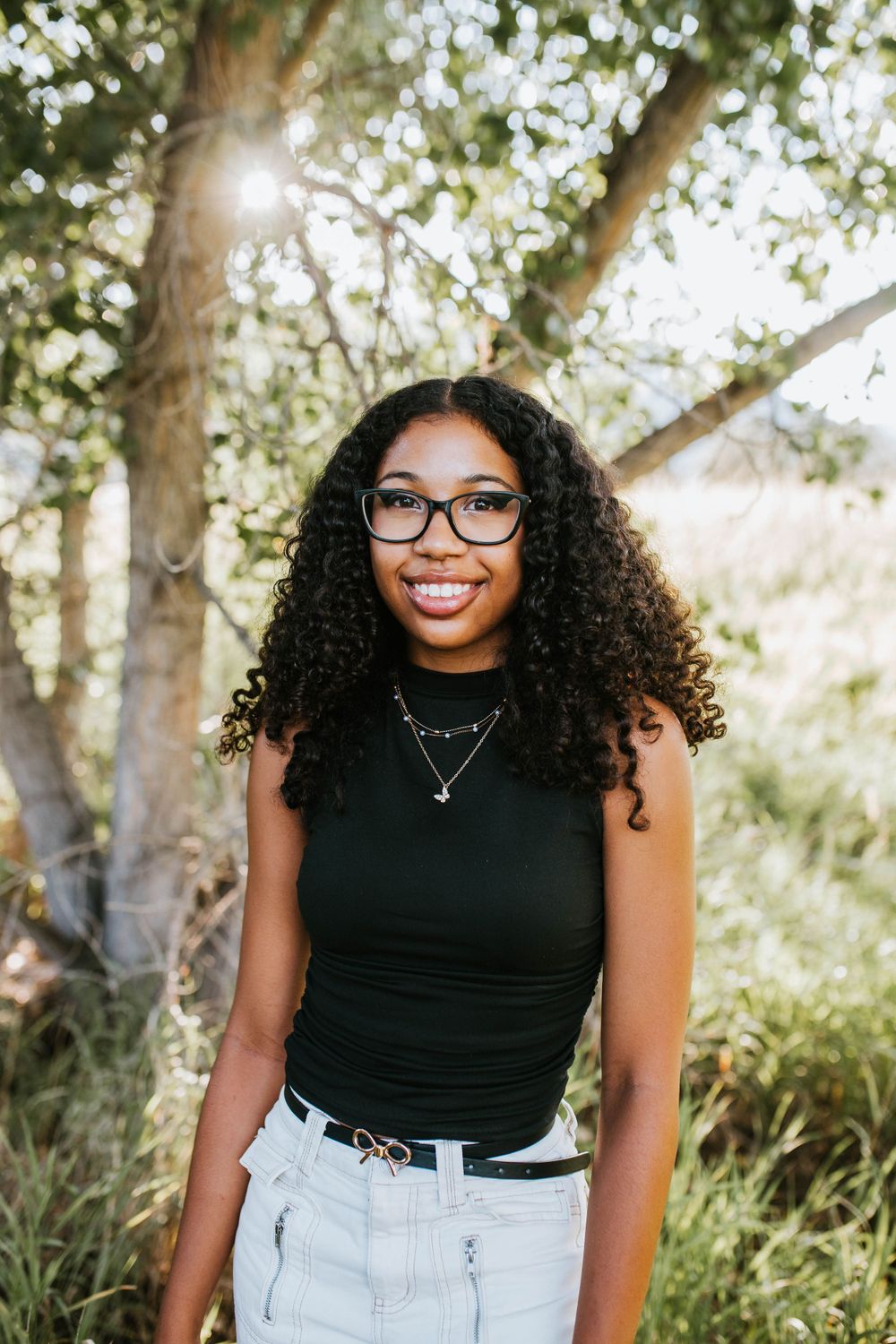 Outdoor portrait of person in black tank top and glasses with natural sunlit trees in background.