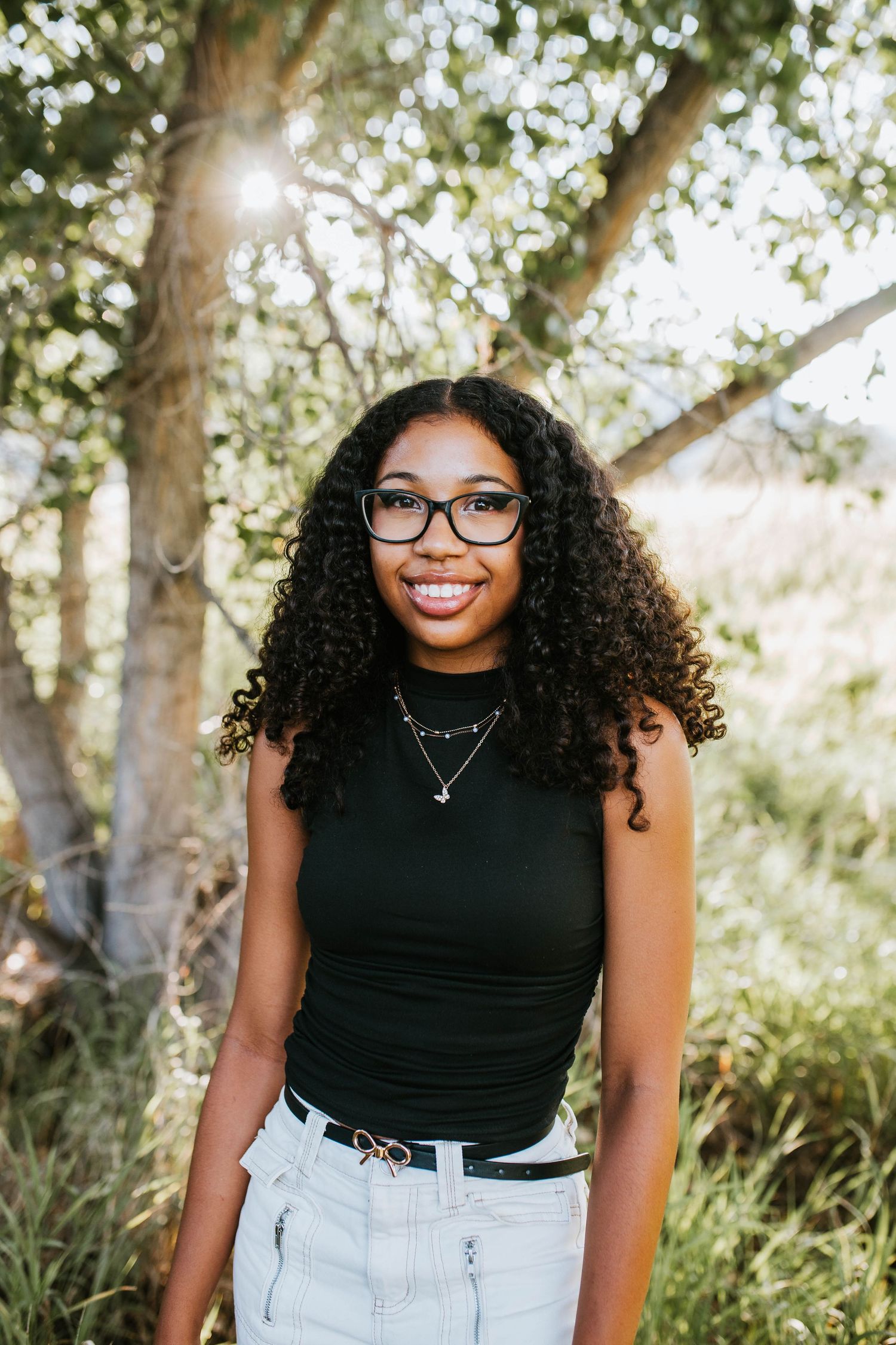 Outdoor portrait of person in black tank top and glasses with natural sunlit trees in background.