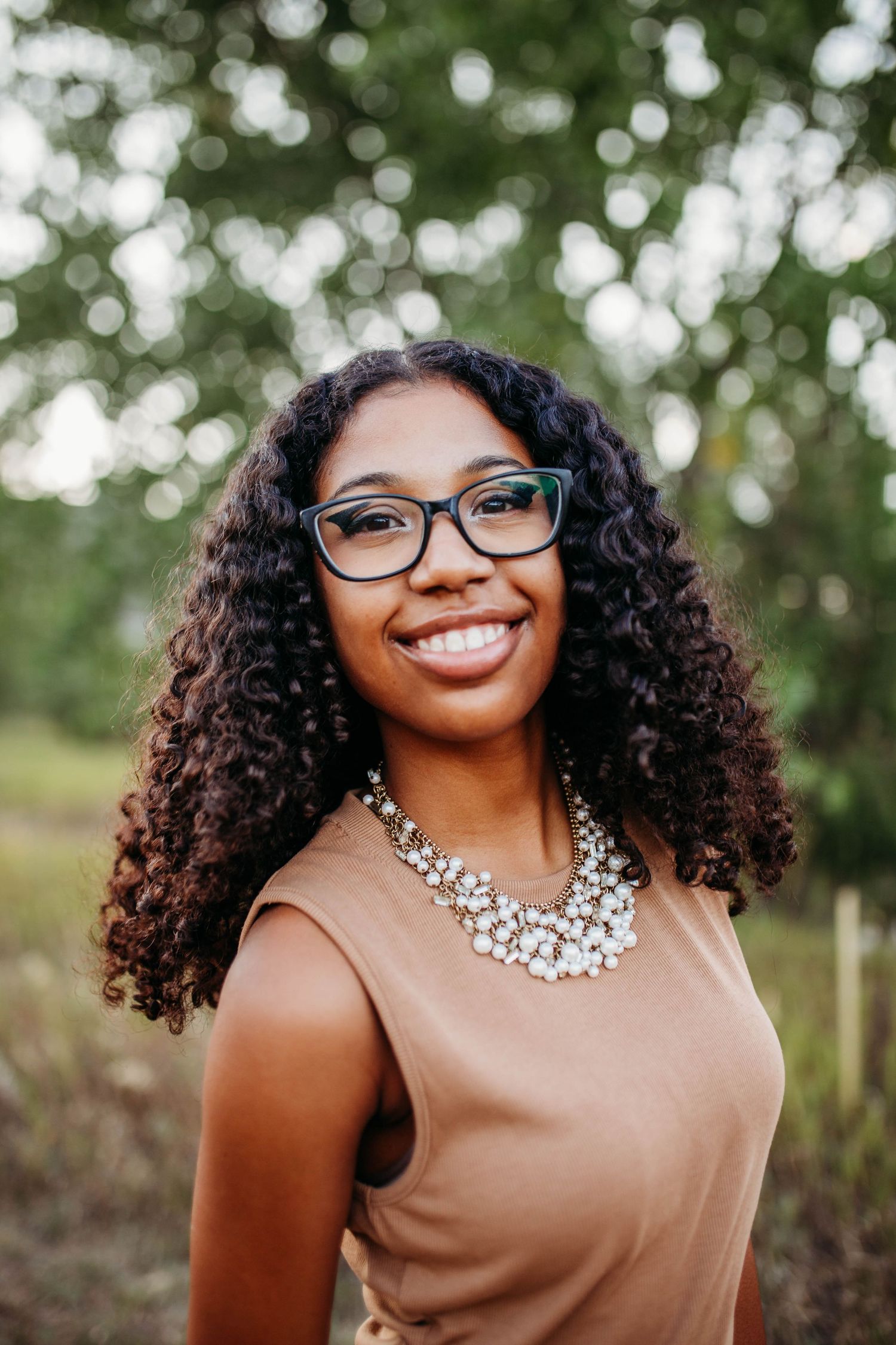 Portrait features person in tan top with statement necklace and glasses against blurred green backdrop.