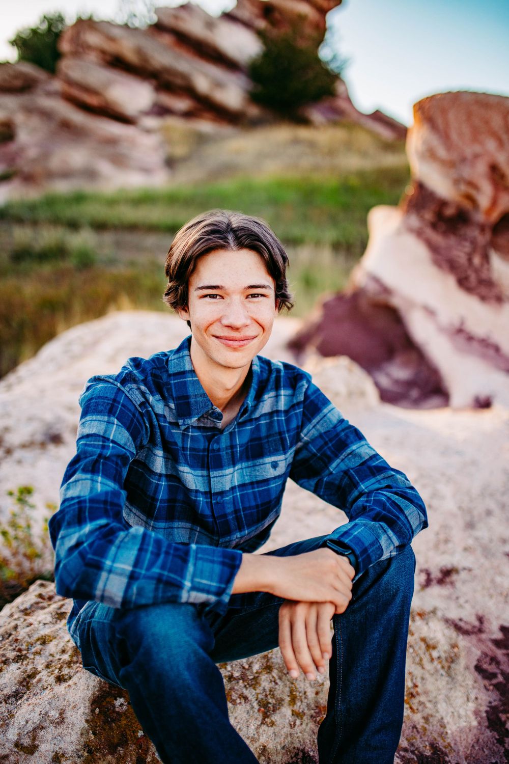 Person in blue plaid shirt sitting on red rocks with natural geological formations in background.