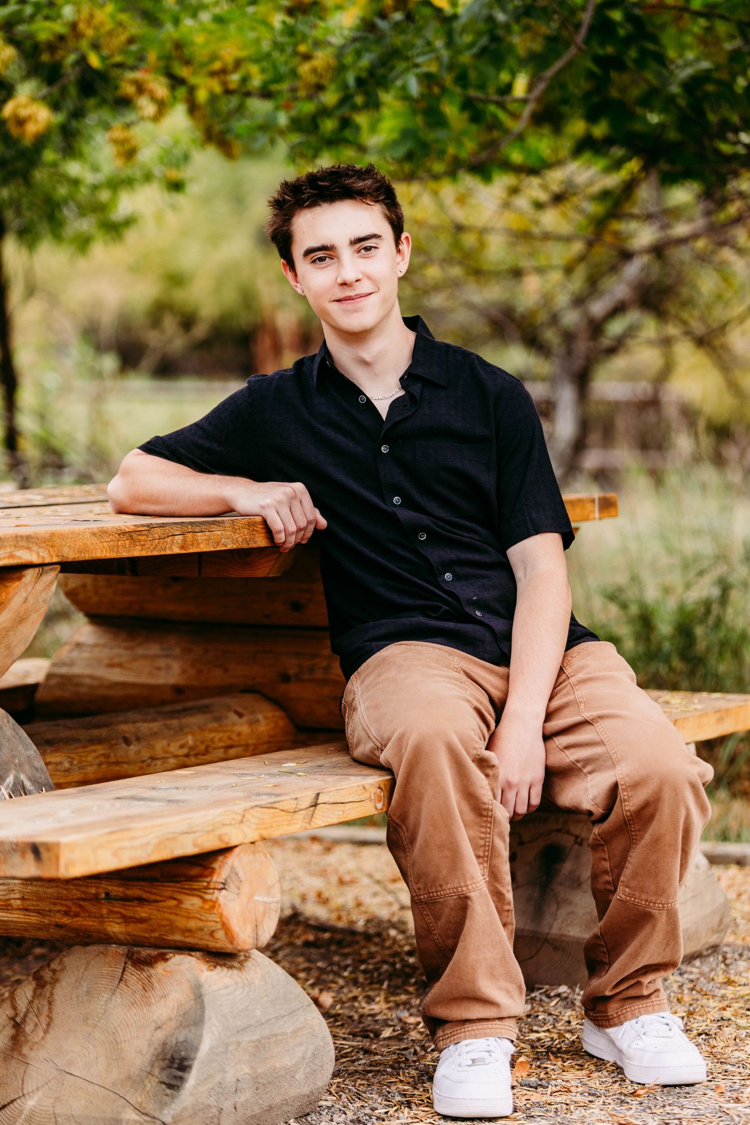 Person in black polo shirt sitting on wooden bench outdoors wearing khaki pants and white shoes.