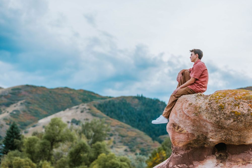 Person sitting on large rock formation overlooking mountain landscape at sunset.