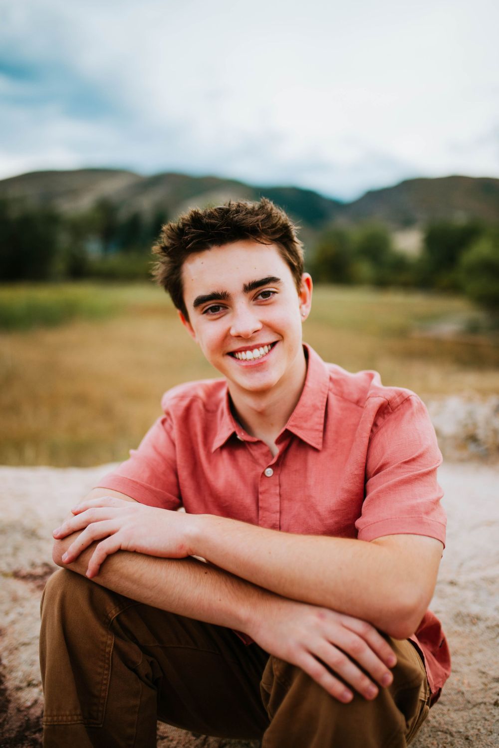 Person in salmon colored polo shirt sitting outdoors against mountain backdrop.