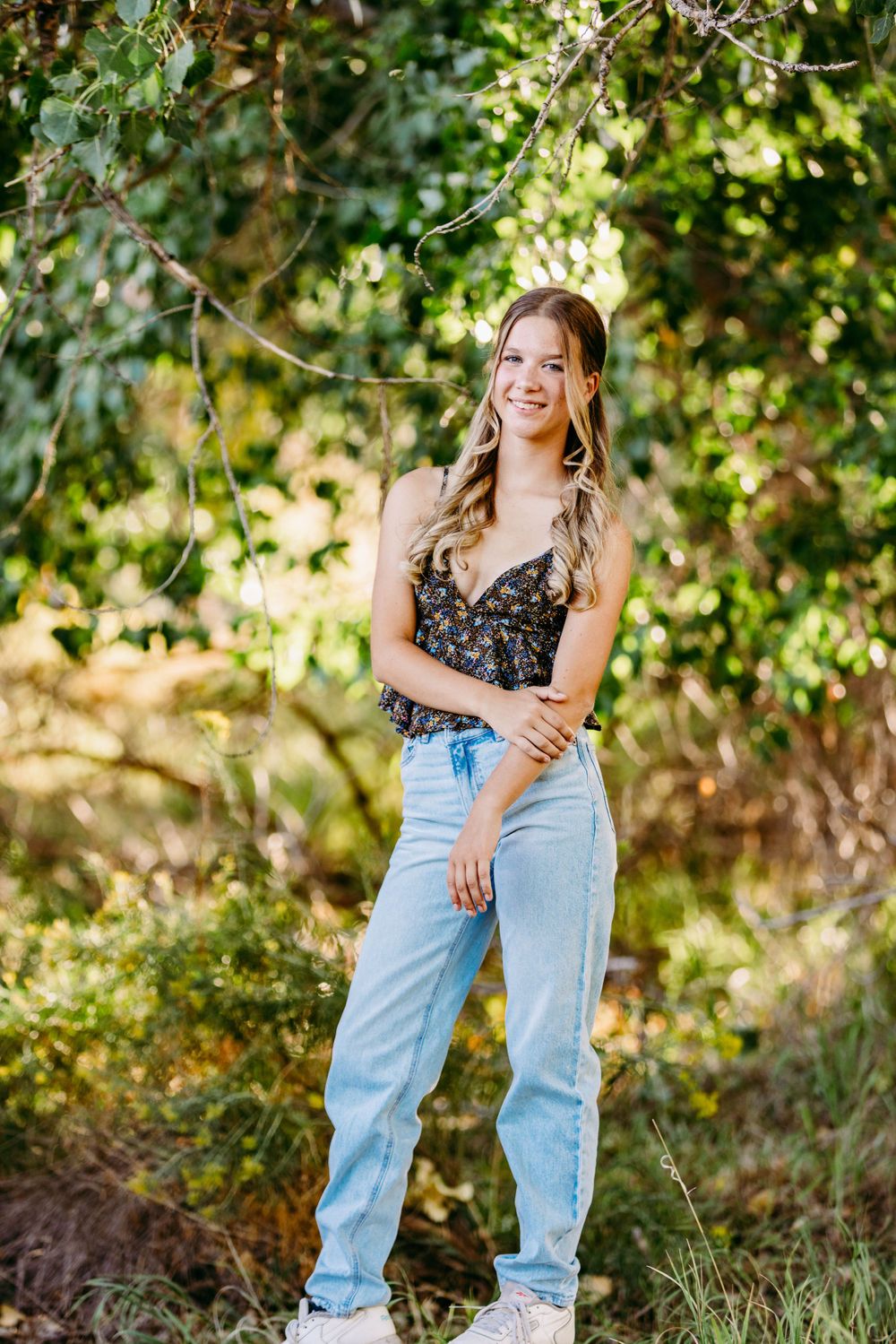 Full length portrait in casual denim and floral top against natural greenery.
