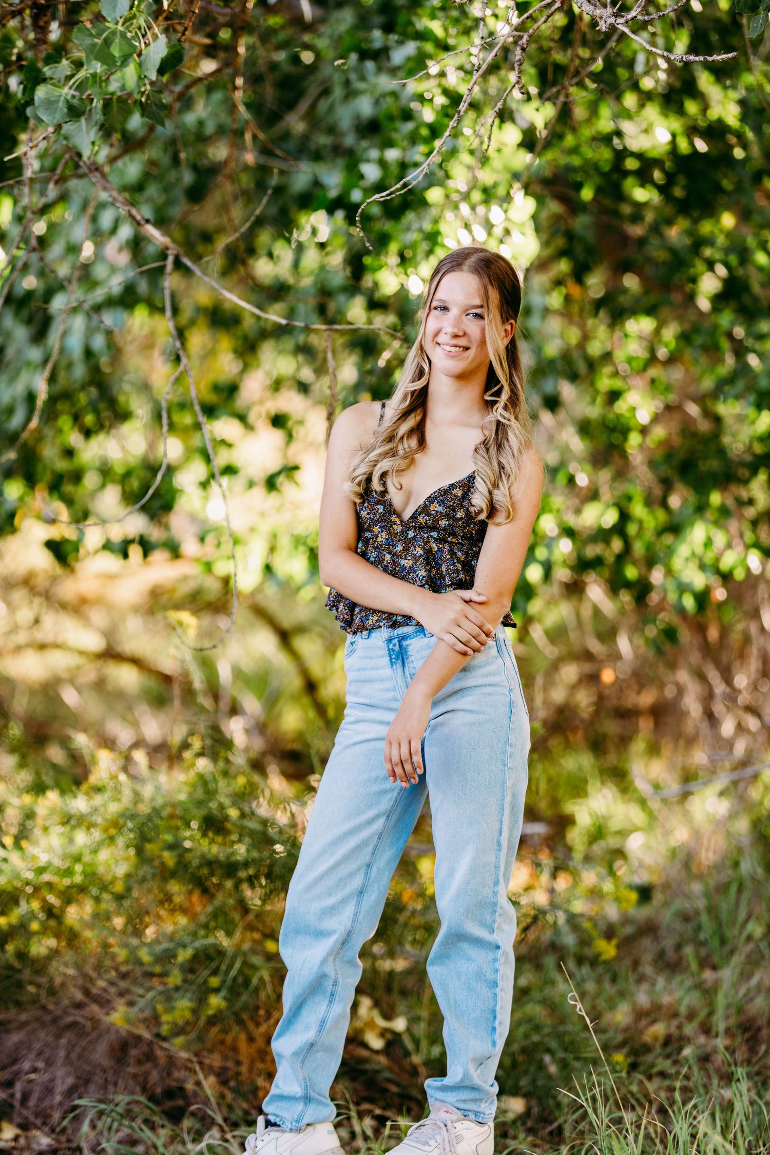 Full length portrait in casual denim and floral top against natural greenery.
