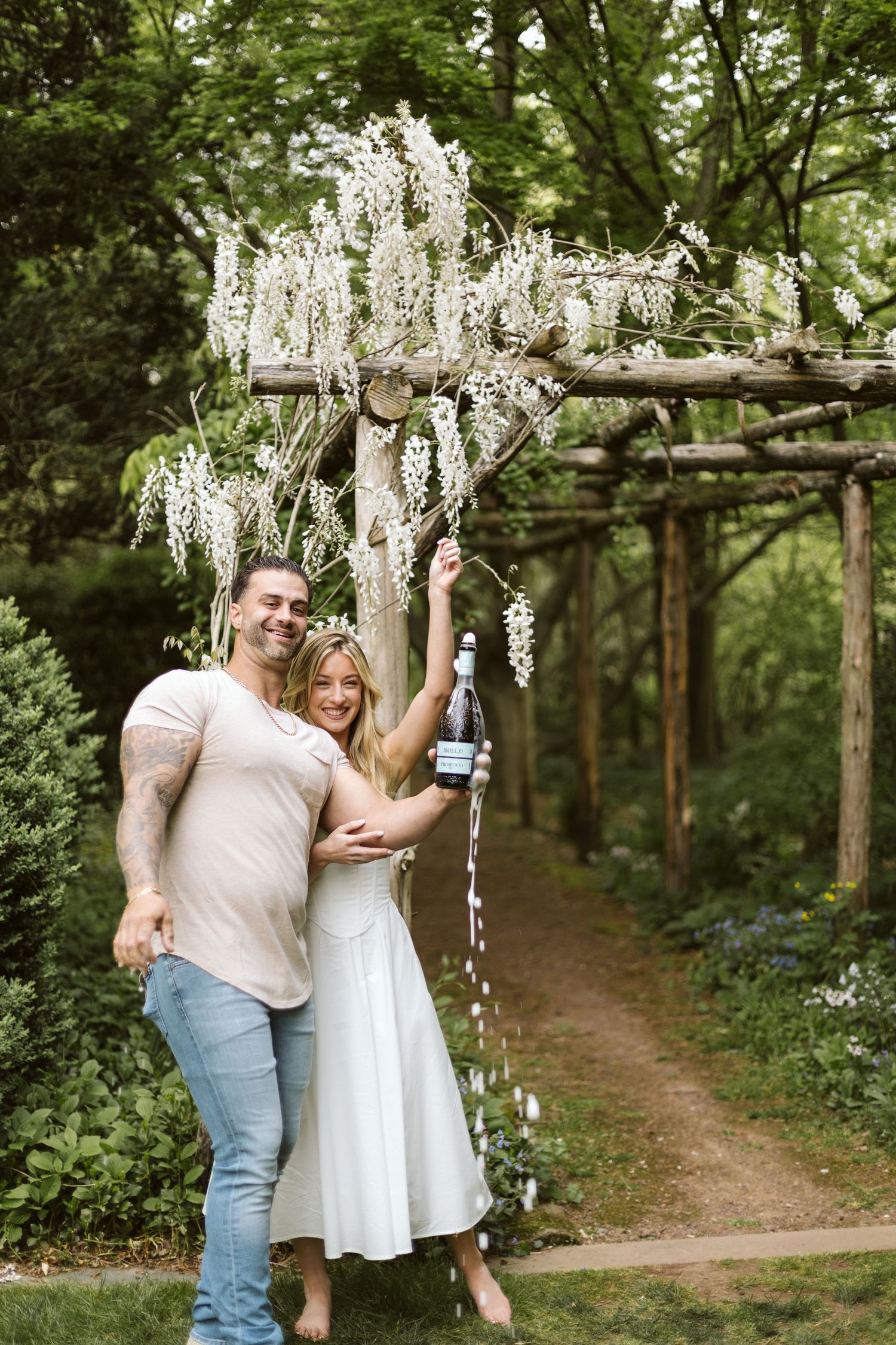 A couple celebrates with champagne under a wisteria-covered garden arbor.