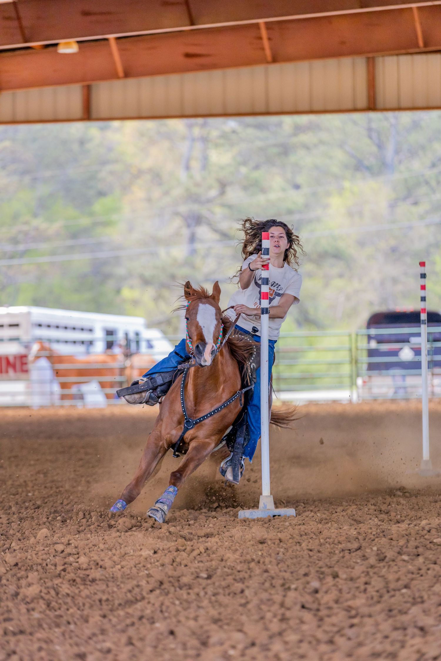 Rider guiding horse through barrel racing competition in dusty indoor arena.