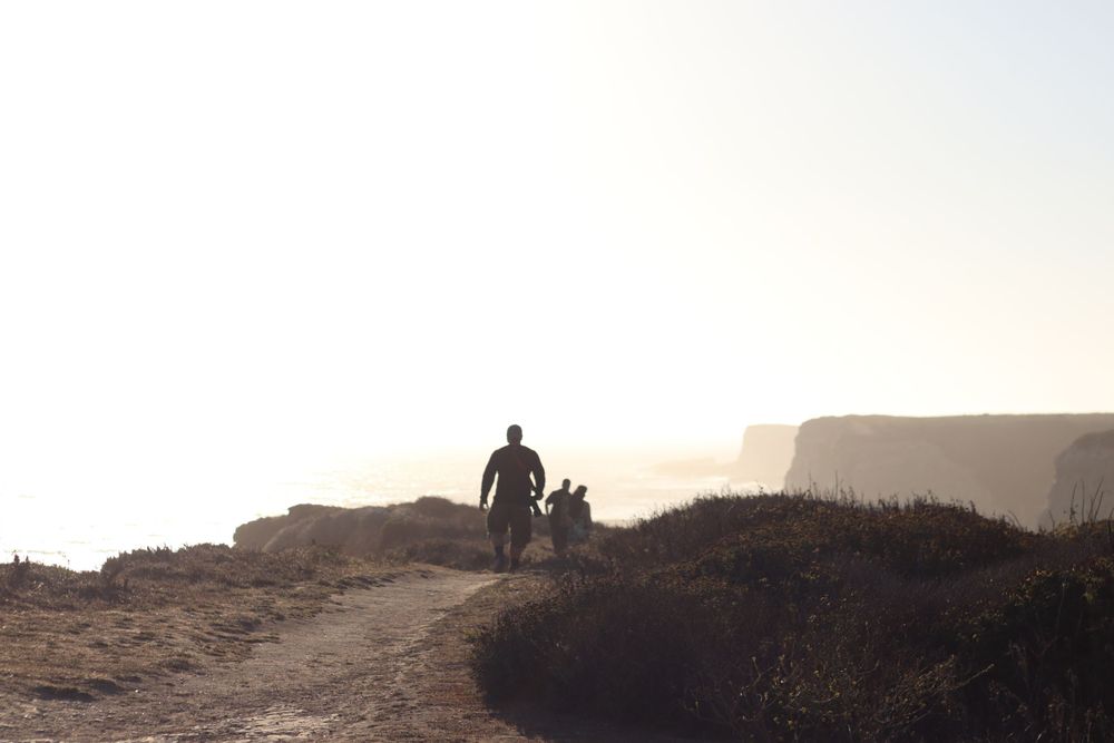 Silhouetted figure walks along a scenic clifftop trail at sunset against a mountainous backdrop.