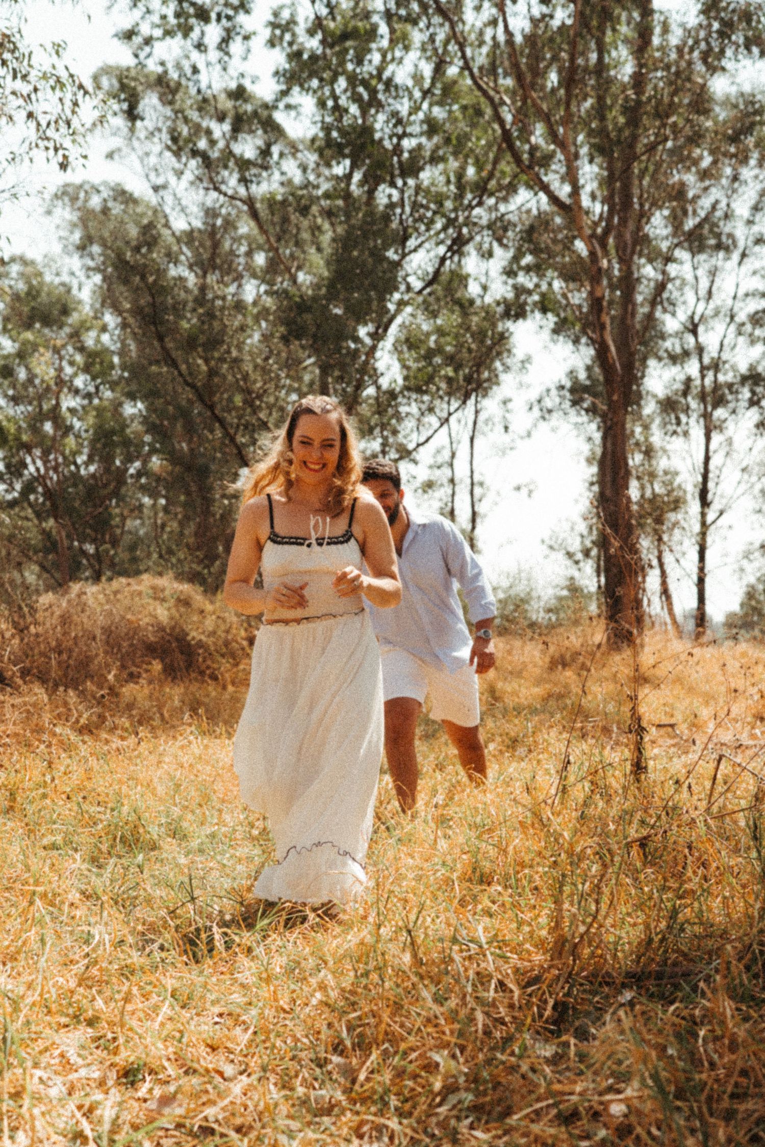A couple runs playfully through a sun-drenched field with eucalyptus trees in the background.