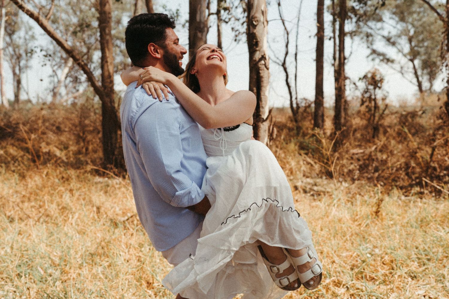 Romantic couple embraces in a sunlit forest setting wearing coordinated white and blue outfits.