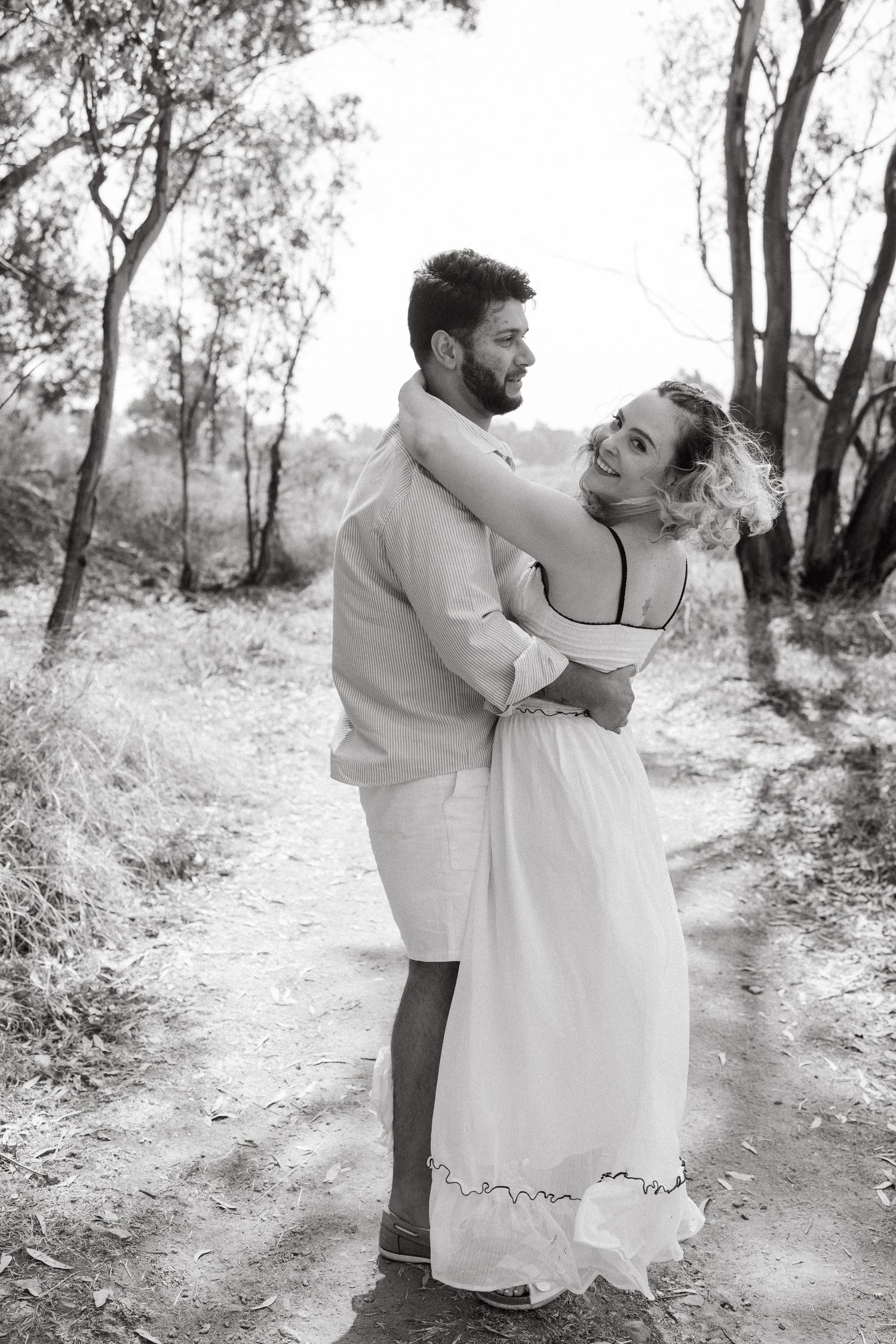 A couple dances together on a rustic dirt path surrounded by trees in black and white.