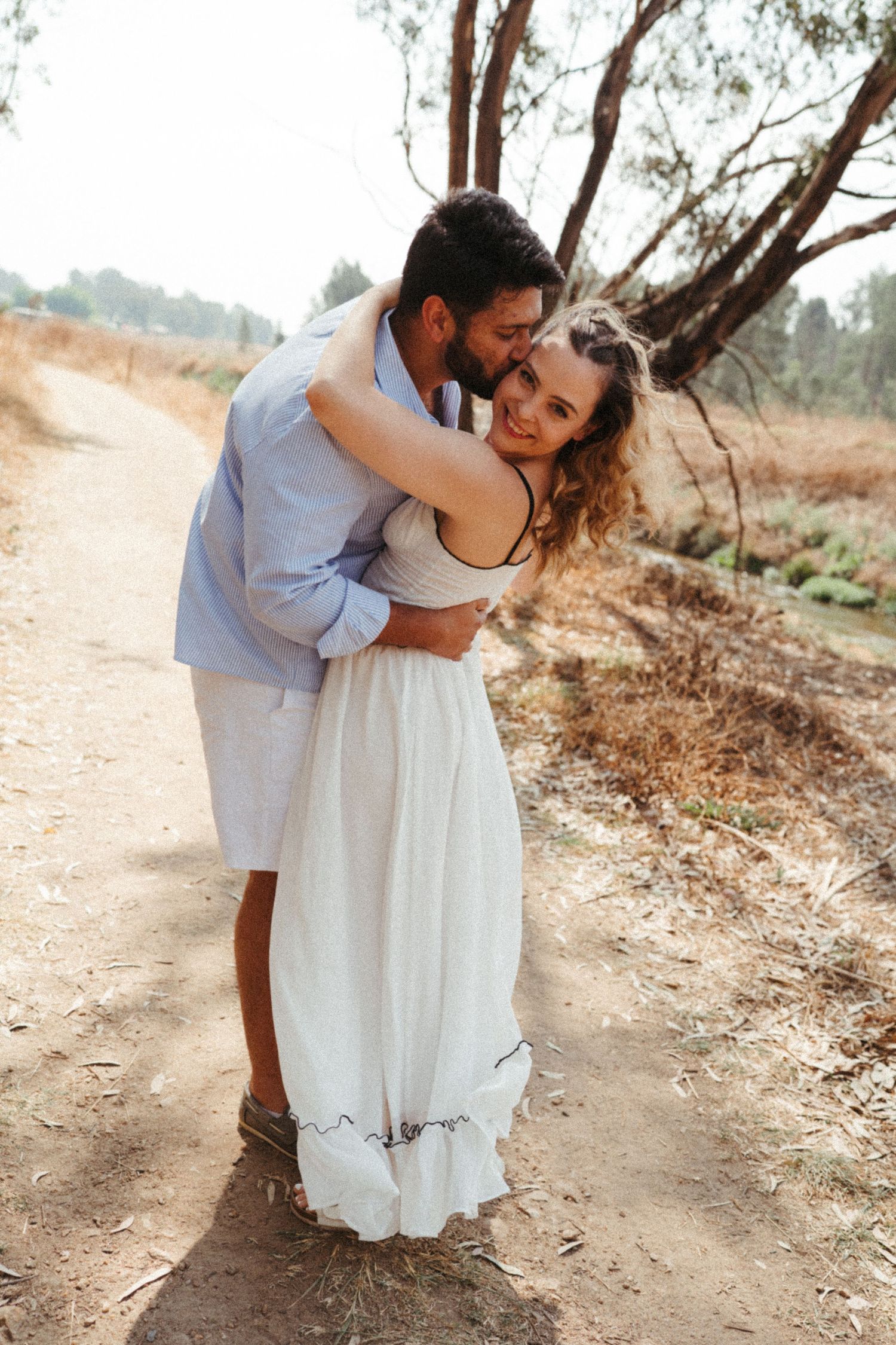 A loving couple embraces on a sunlit nature trail wearing coordinated white outfits.