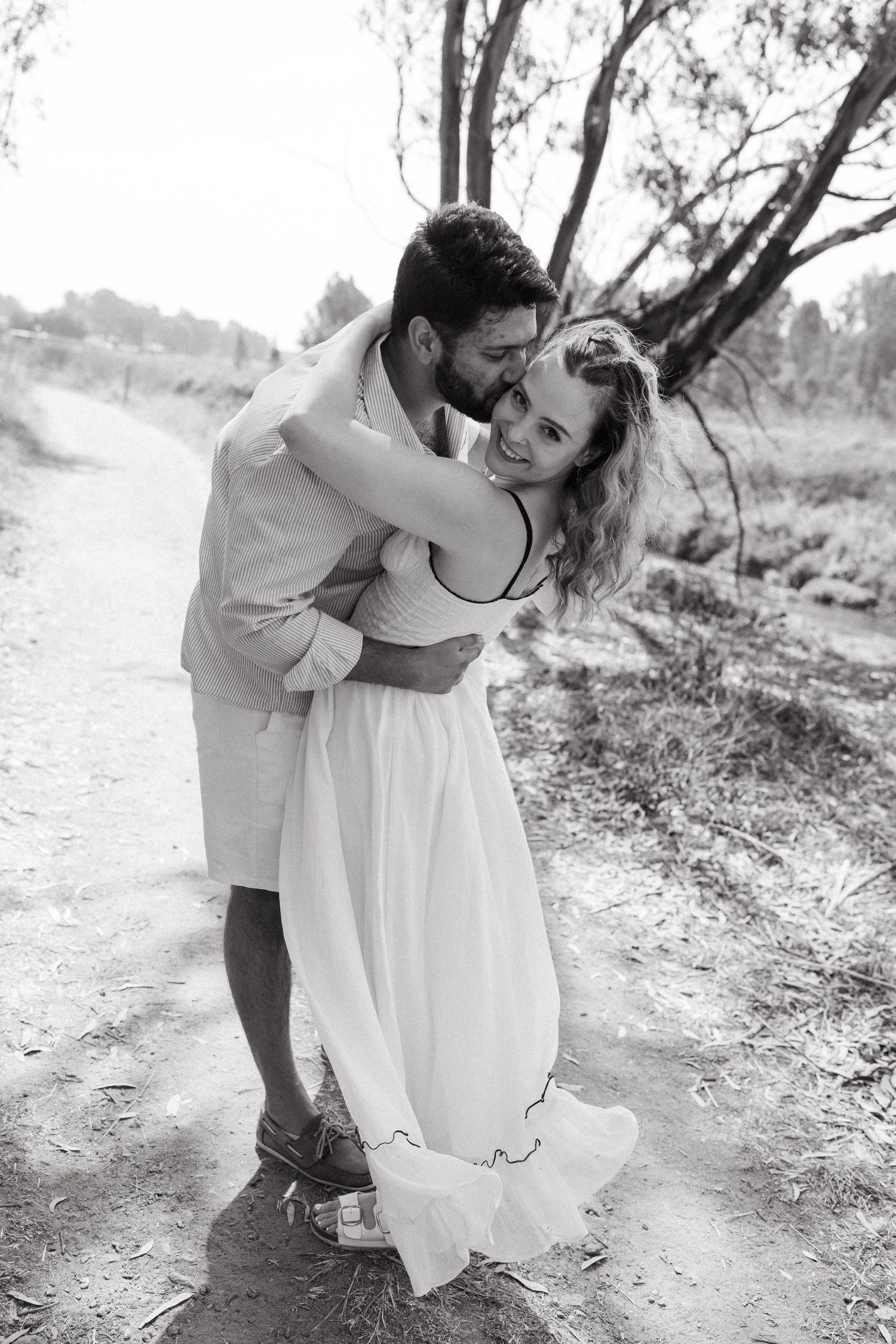 A couple embraces in a romantic pose on a dirt path surrounded by trees.