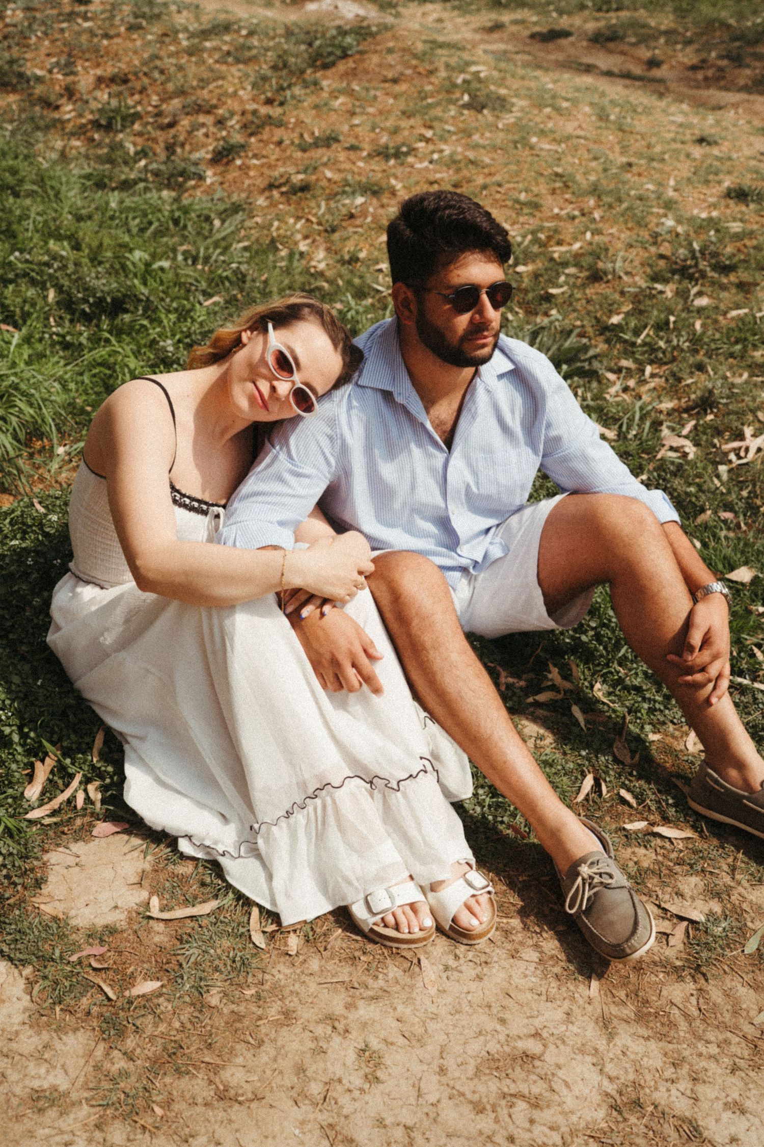 A relaxed couple sits together on the ground in coordinated summer outfits.