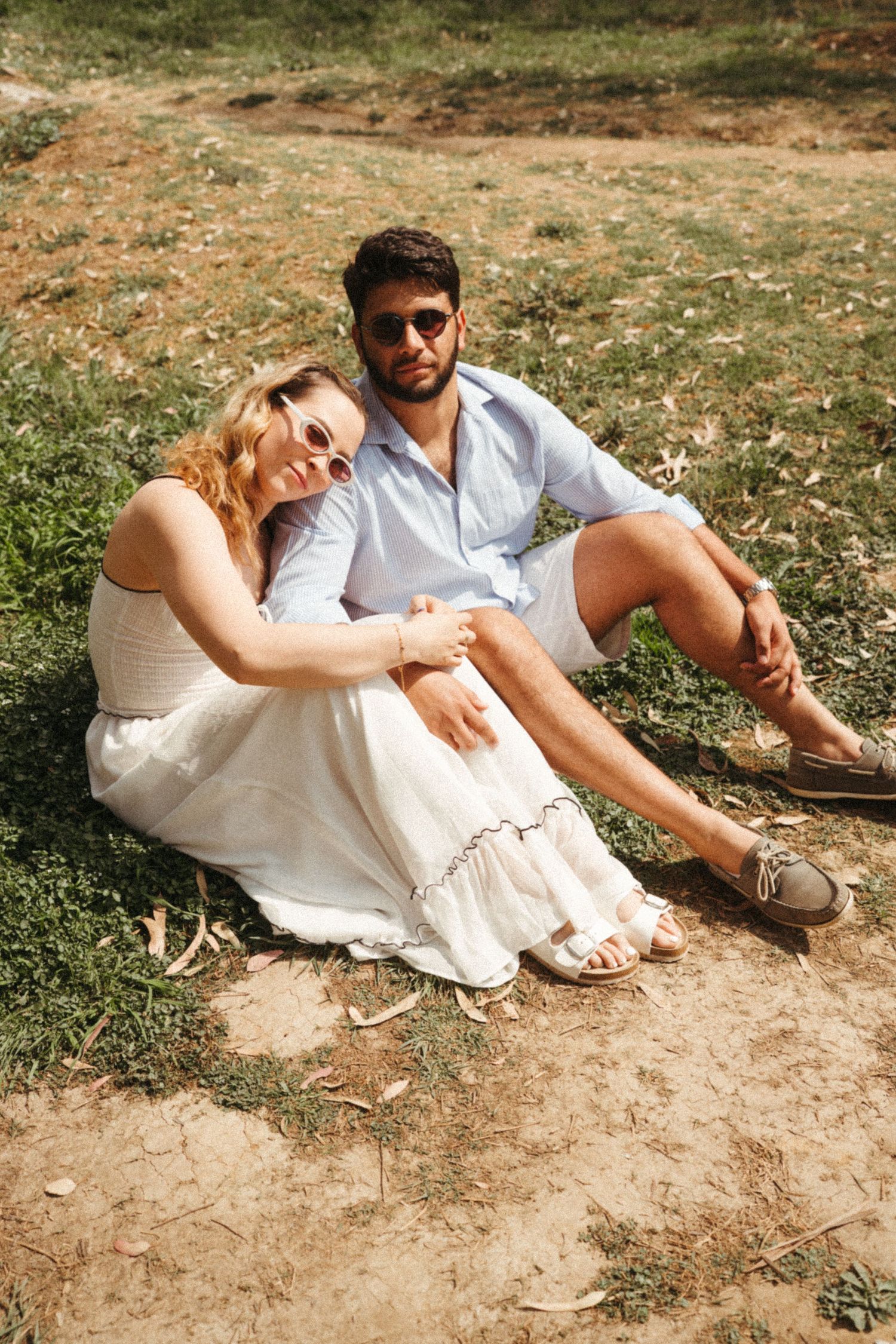 A couple in white summer outfits relaxes together on grassy ground.