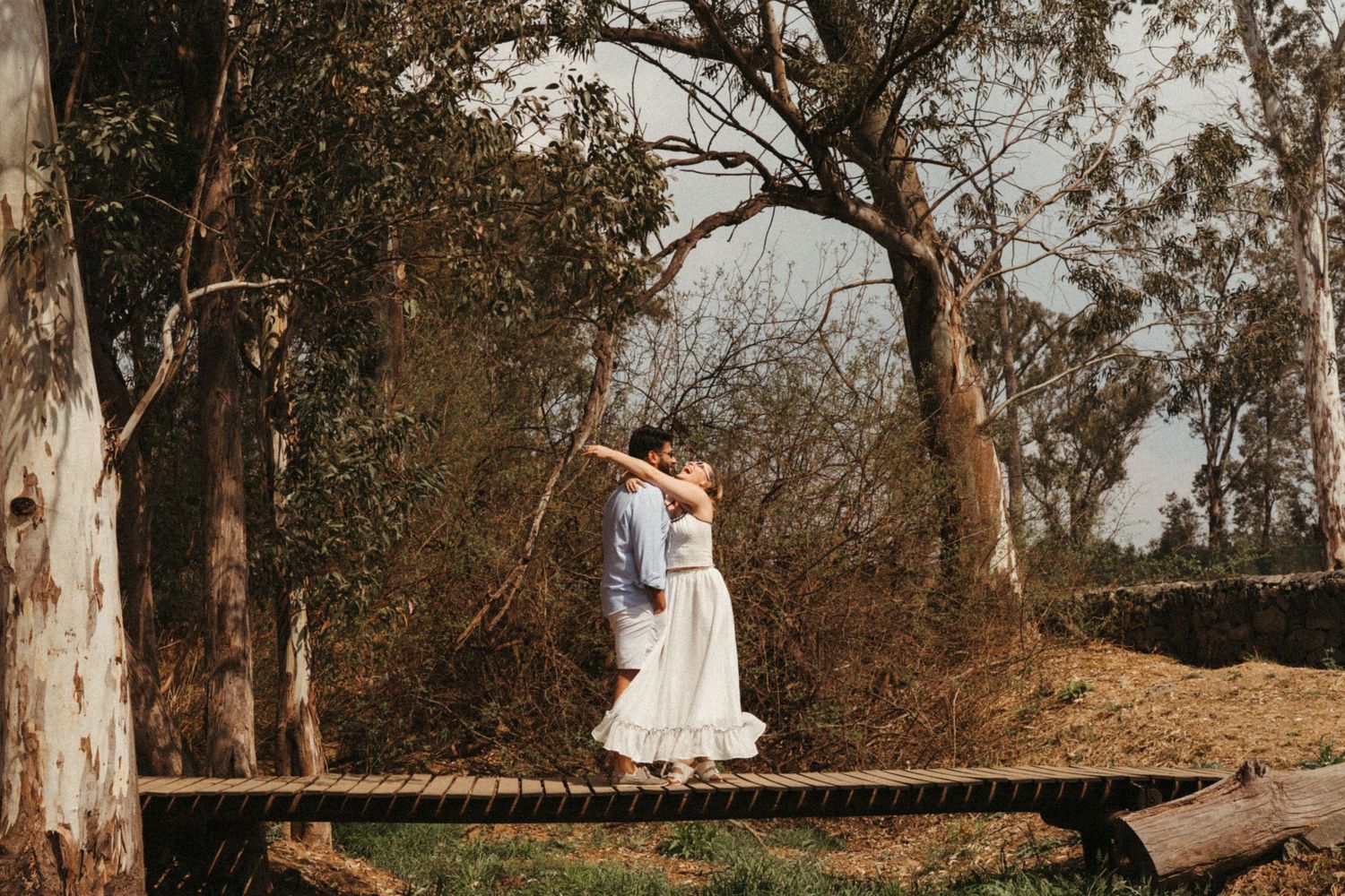 Romantic couple embracing on a wooden bridge surrounded by tall trees.
